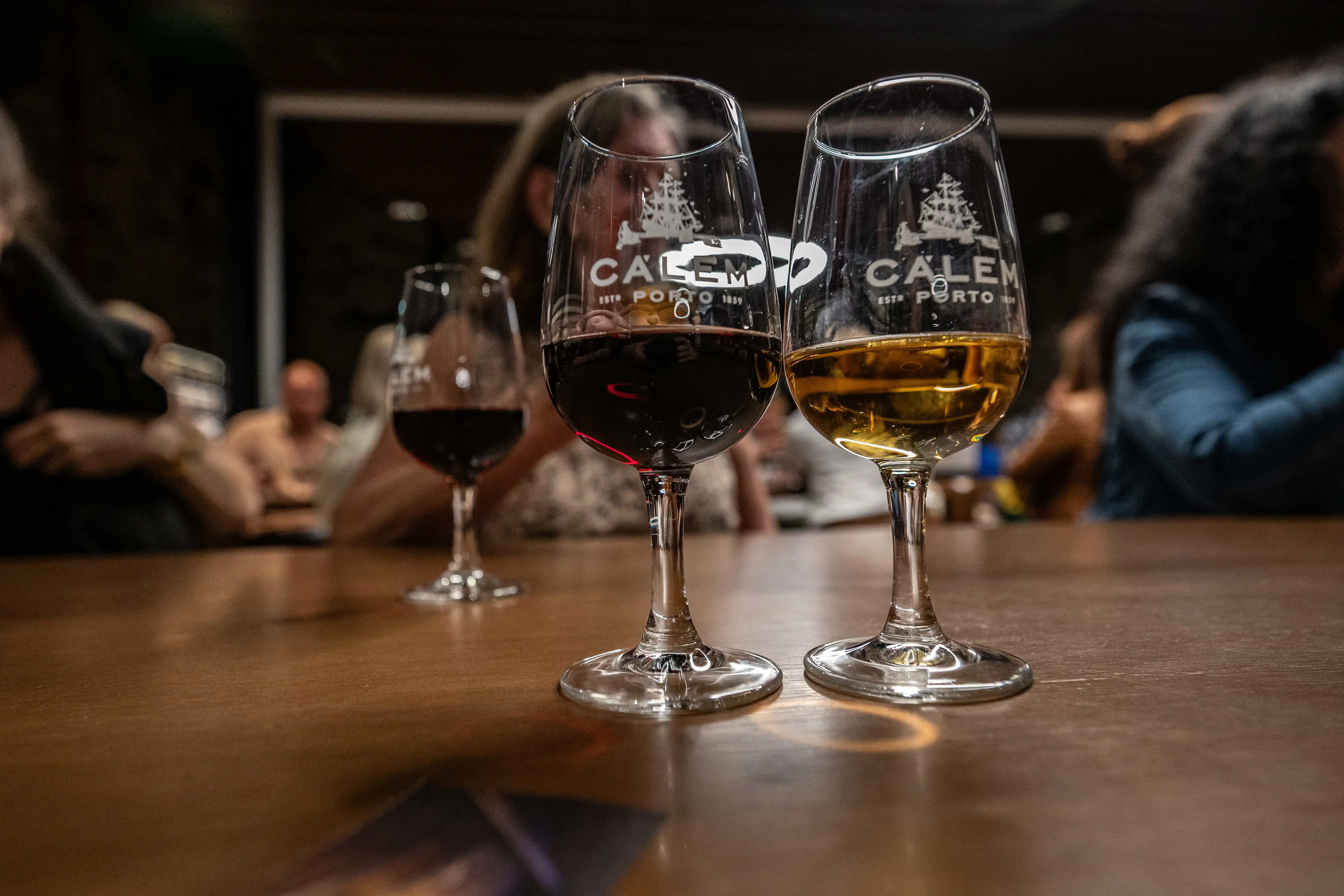 Two Calem Port wine glasses, one with dark red port and the other with amber-coloured port, sit on a wooden table. The Calem logo, featuring a ship, is visible on both glasses. In the softly blurred background, other people are present, suggesting a tasting or social gathering.