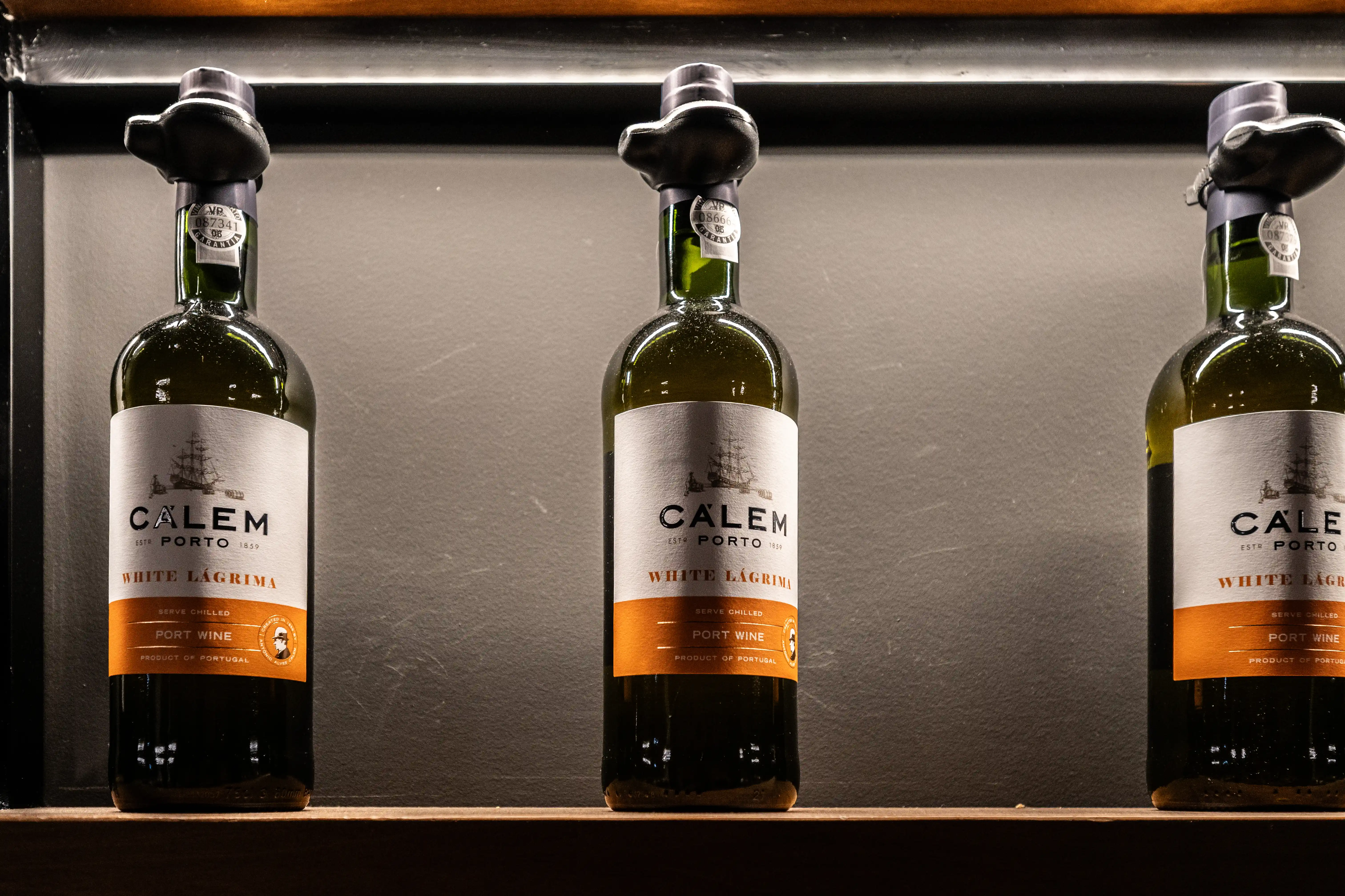 Three green glass bottles of Calem White Lágrica Port Wine are neatly arranged on a wooden shelf. Each bottle has a white label with black text detailing the brand CALEM PORTO and the wine type WHITE LÁGRIMA, alongside an orange band stating PORT WINE and PRODUCT OF PORTUGAL. The bottles are capped with dark grey closures.