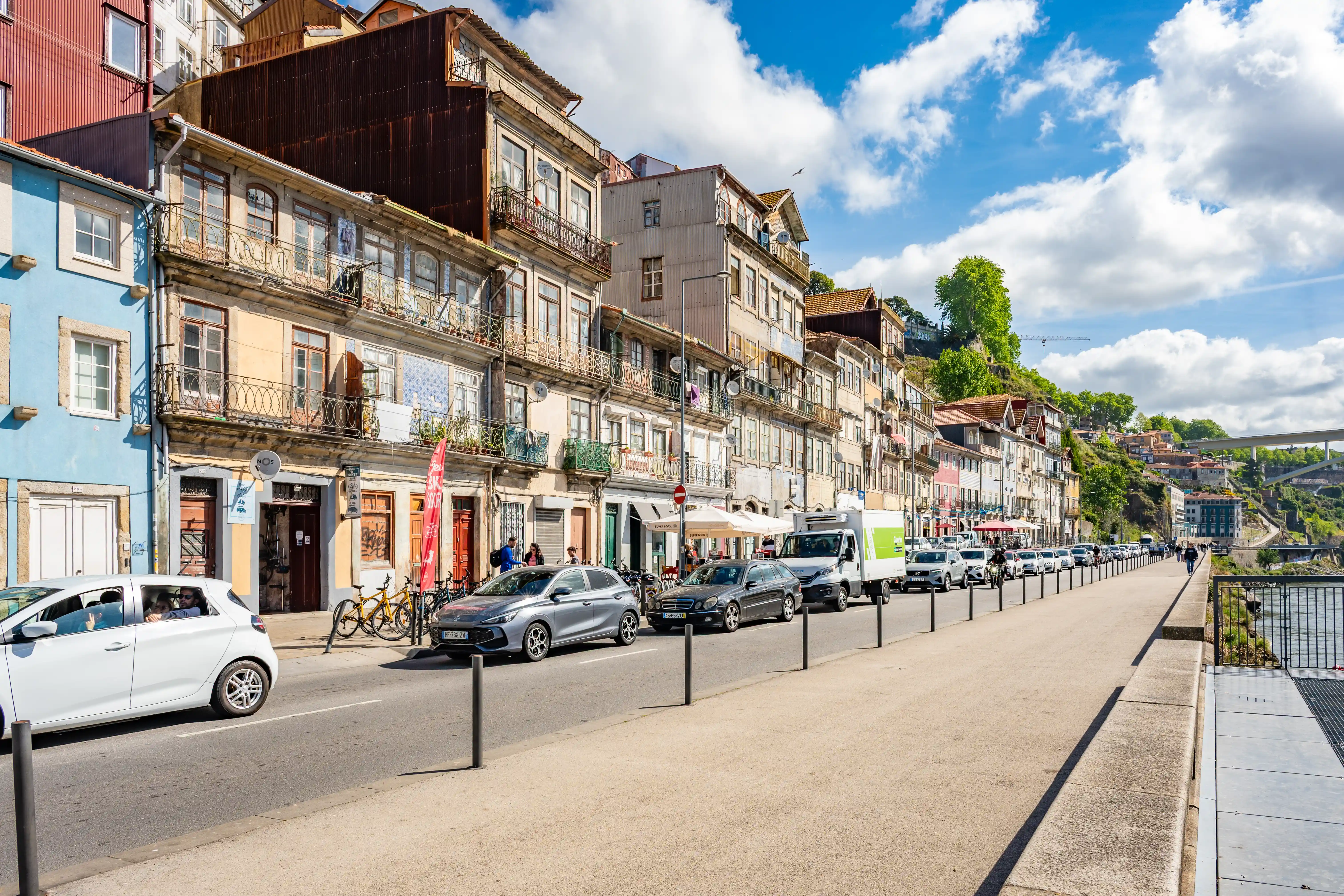 A street view of Porto, Portugal, with historic buildings, a river, a promenade, cars, and a bridge under a blue sky with white clouds.