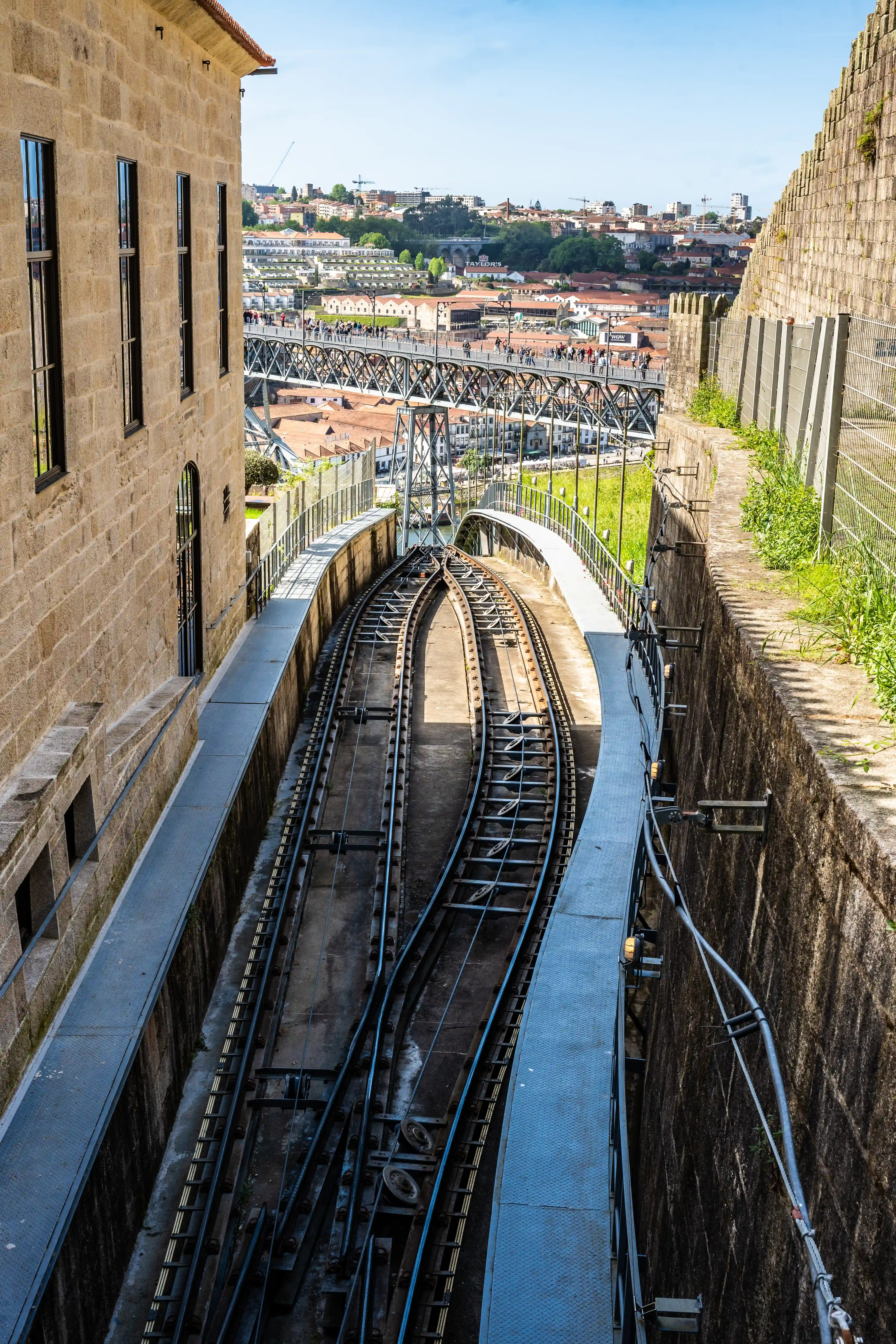 Steep funicular railway tracks descend between stone walls, with the Dom Luís I Bridge and the city of Porto visible in the background under a blue sky.