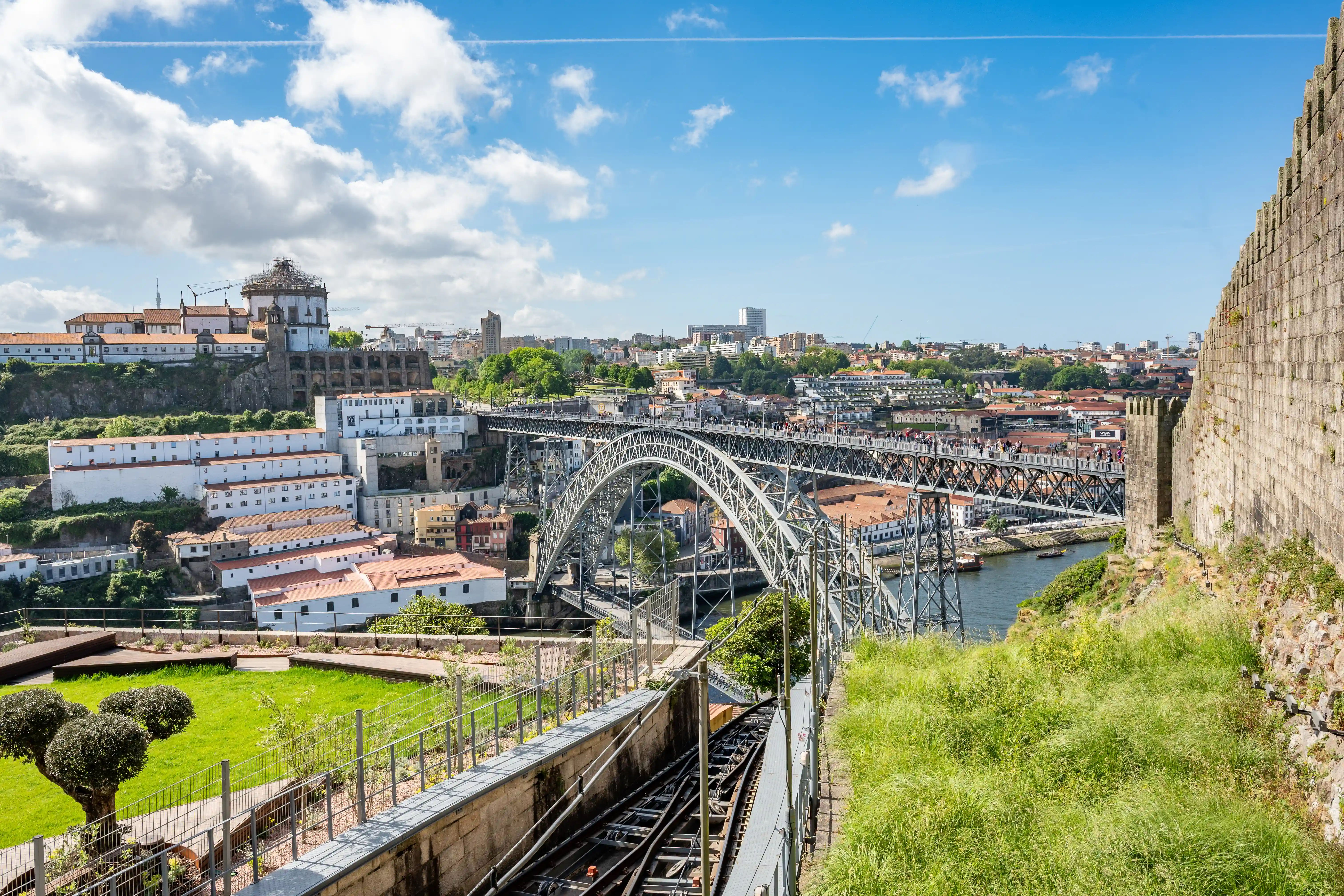 A view of the Dom Luís I Bridge in Porto, Portugal, from a viewpoint overlooking the Douro River, with the Ribeira district and a funicular railway in the foreground.