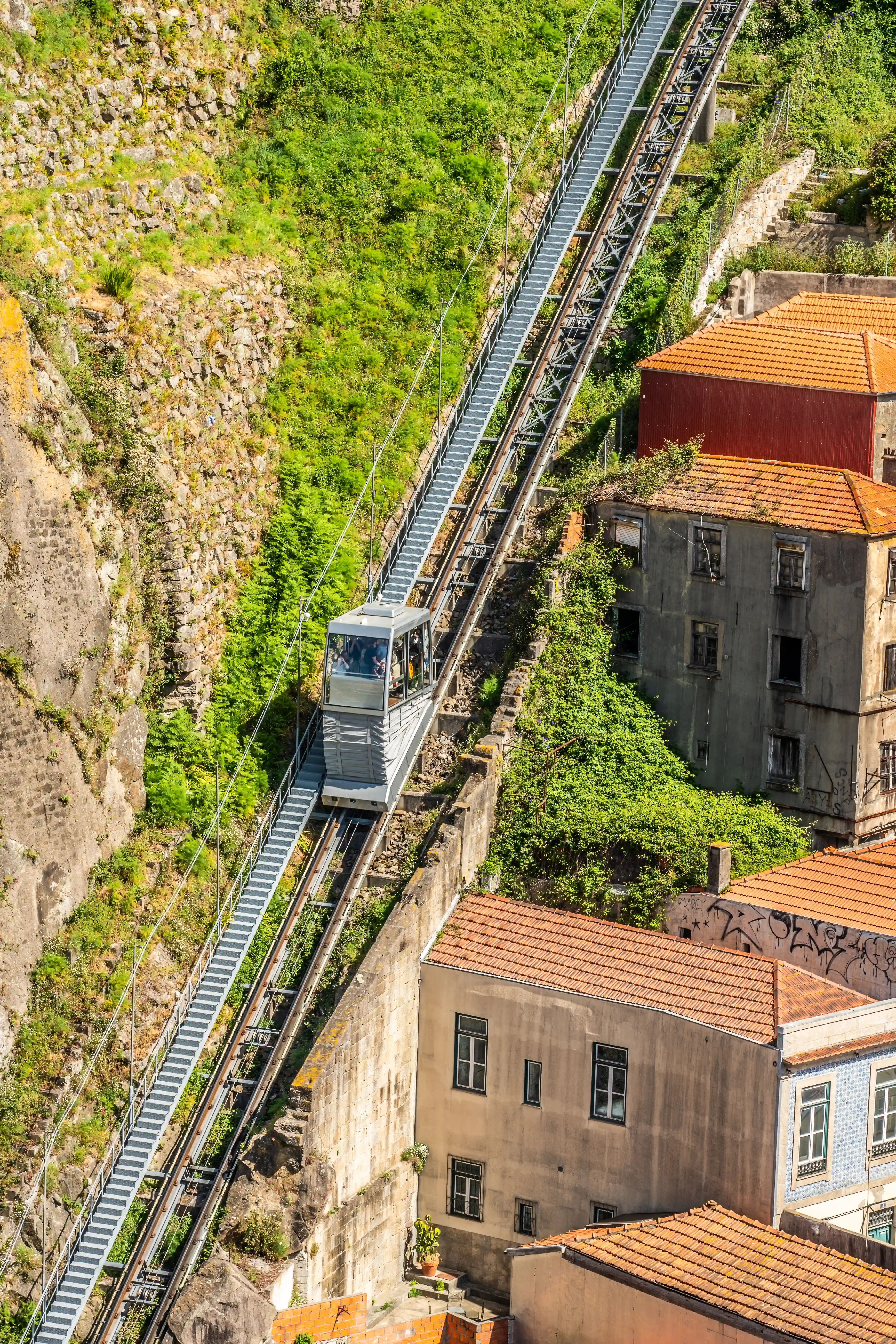 A grey funicular carriage is ascending a steep metal railway track on a sunny day, with lush green vegetation and rocky terrain on one side and old buildings with orange tiled roofs on the other.