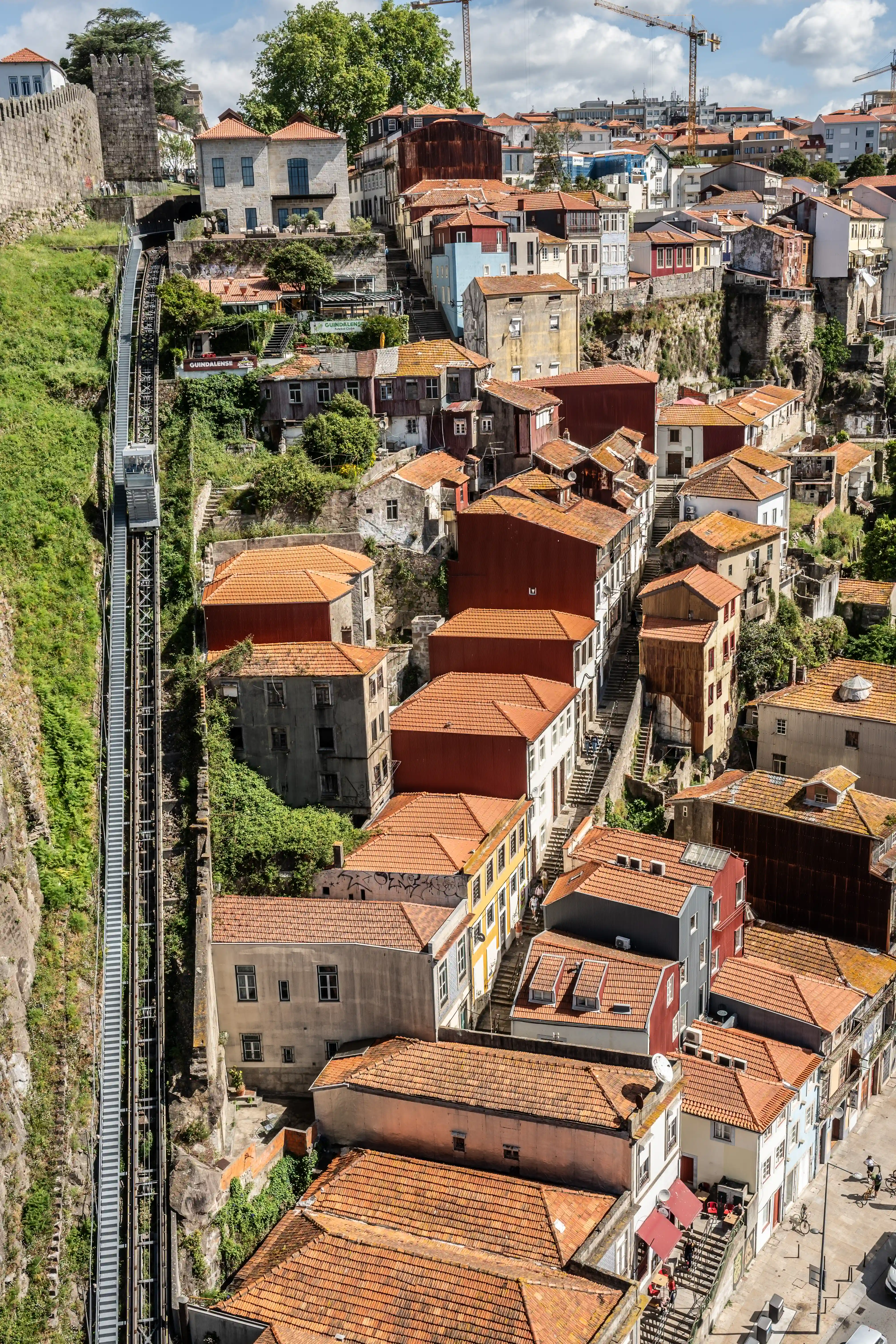 A vertical shot captures a steep hillside in Porto, Portugal, covered in traditional terracotta-tiled buildings. A metal funicular railway runs parallel to a steep, green slope and a stone fortification wall. In the distance, construction cranes are visible above the densely packed house