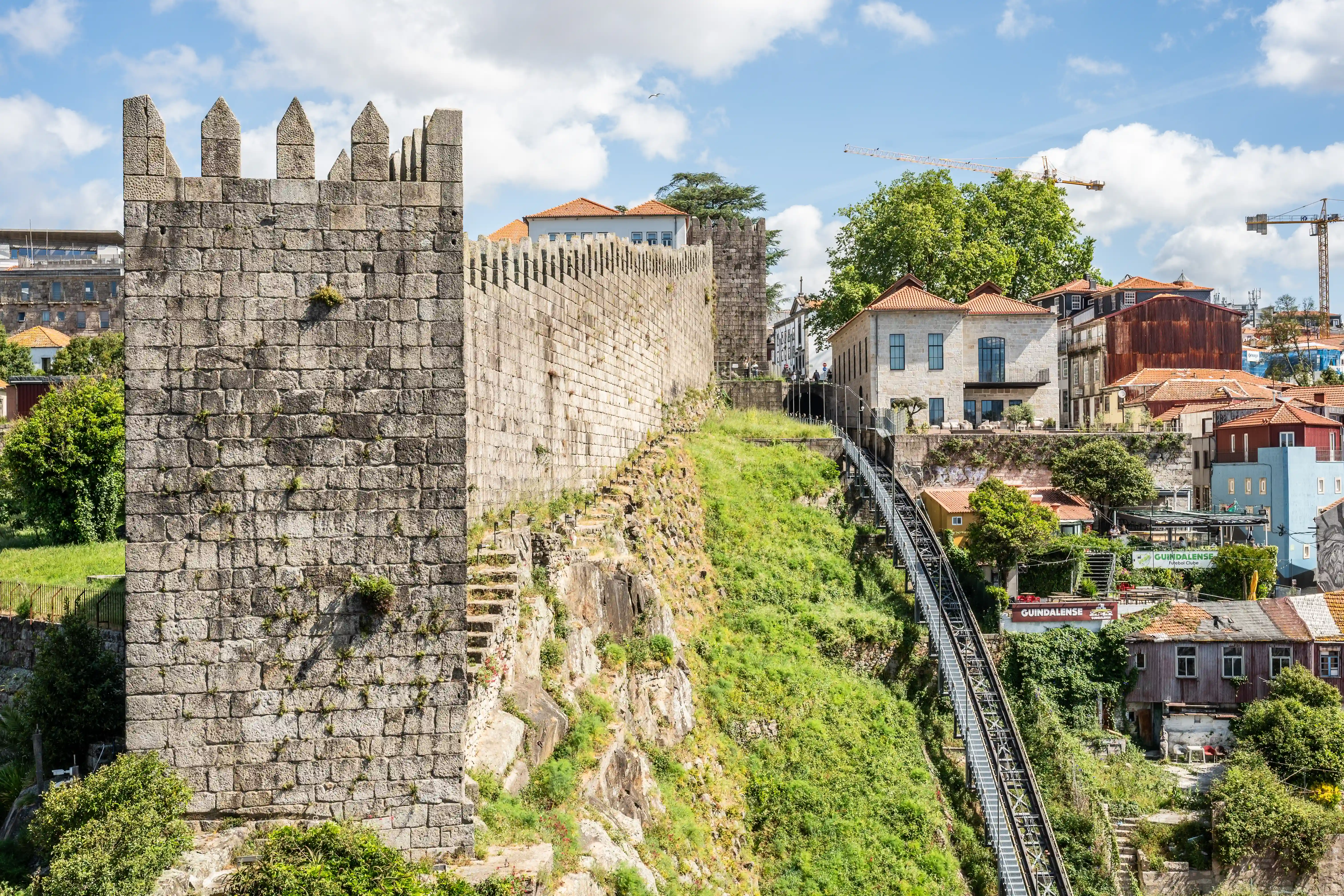 A weathered stone medieval wall with battlements dominates the left side of the frame, overlooking a steep, grassy incline. A metal funicular track zigzags up the hill on the right, bordered by traditional Portuguese buildings with red-tiled roofs. The sky is bright blue with scattered white clouds.