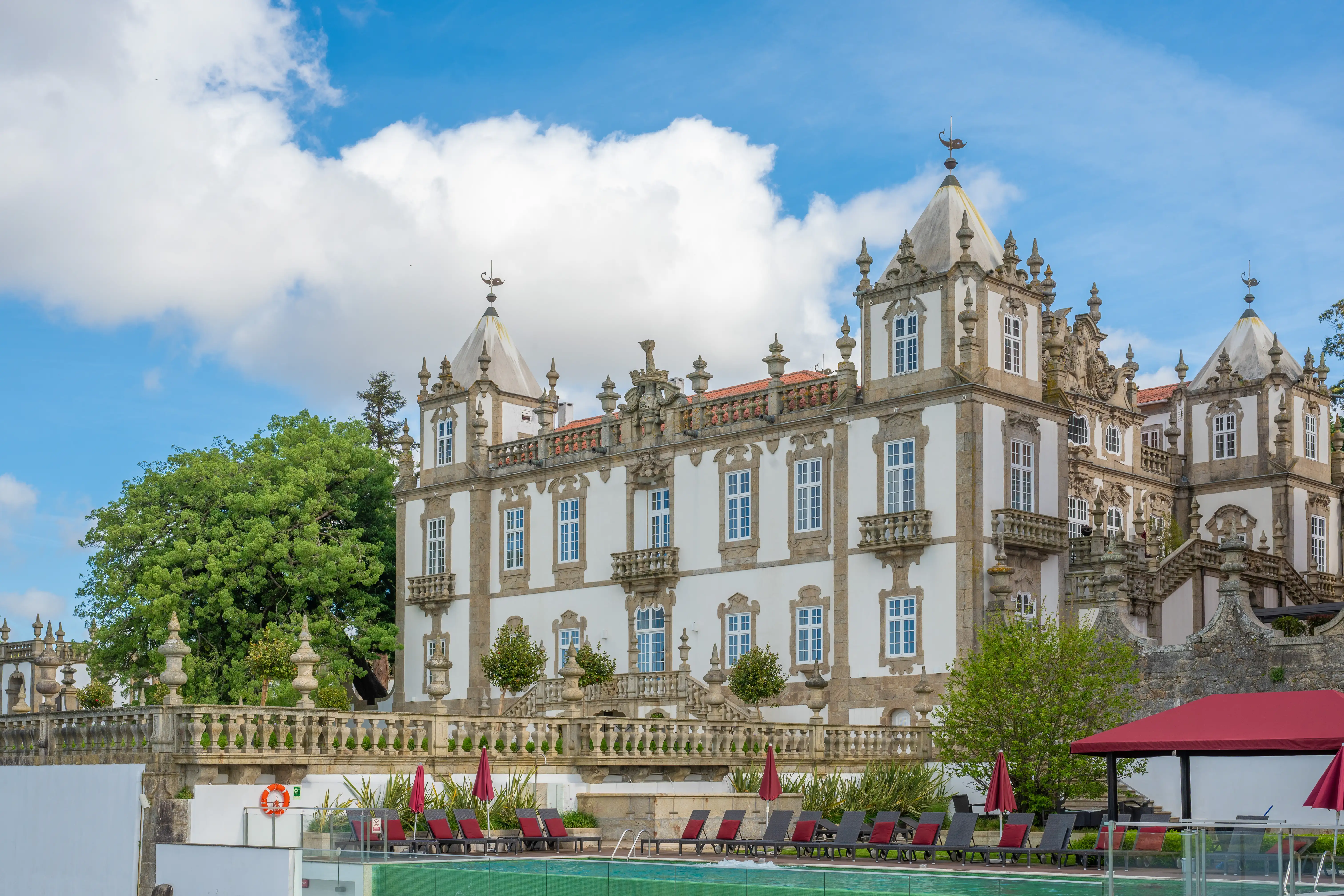 A grand white manor house with ornate white stonework and many windows, featuring decorative turrets topped with weather vanes. A stone balustrade with urns runs across the front of the building, overlooking a swimming pool with red sun loungers and umbrellas.