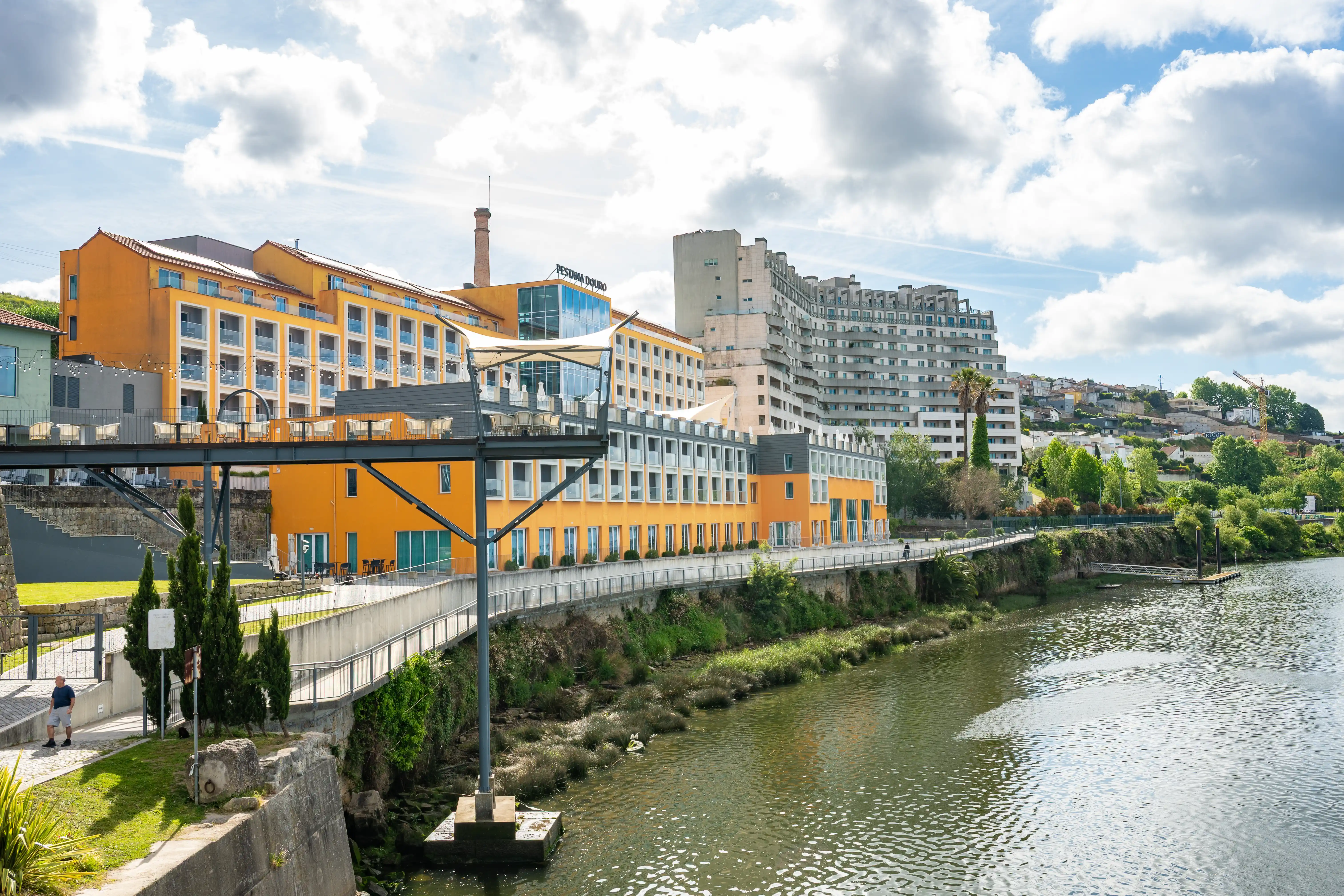 A wide shot of the Douro River in Porto, Portugal, with the vibrant orange Pestana Douro Hotel situated on the riverbank. A modern walkway extends from the hotel over the water, and further buildings and greenery rise up the hill in the background under a cloudy sky.