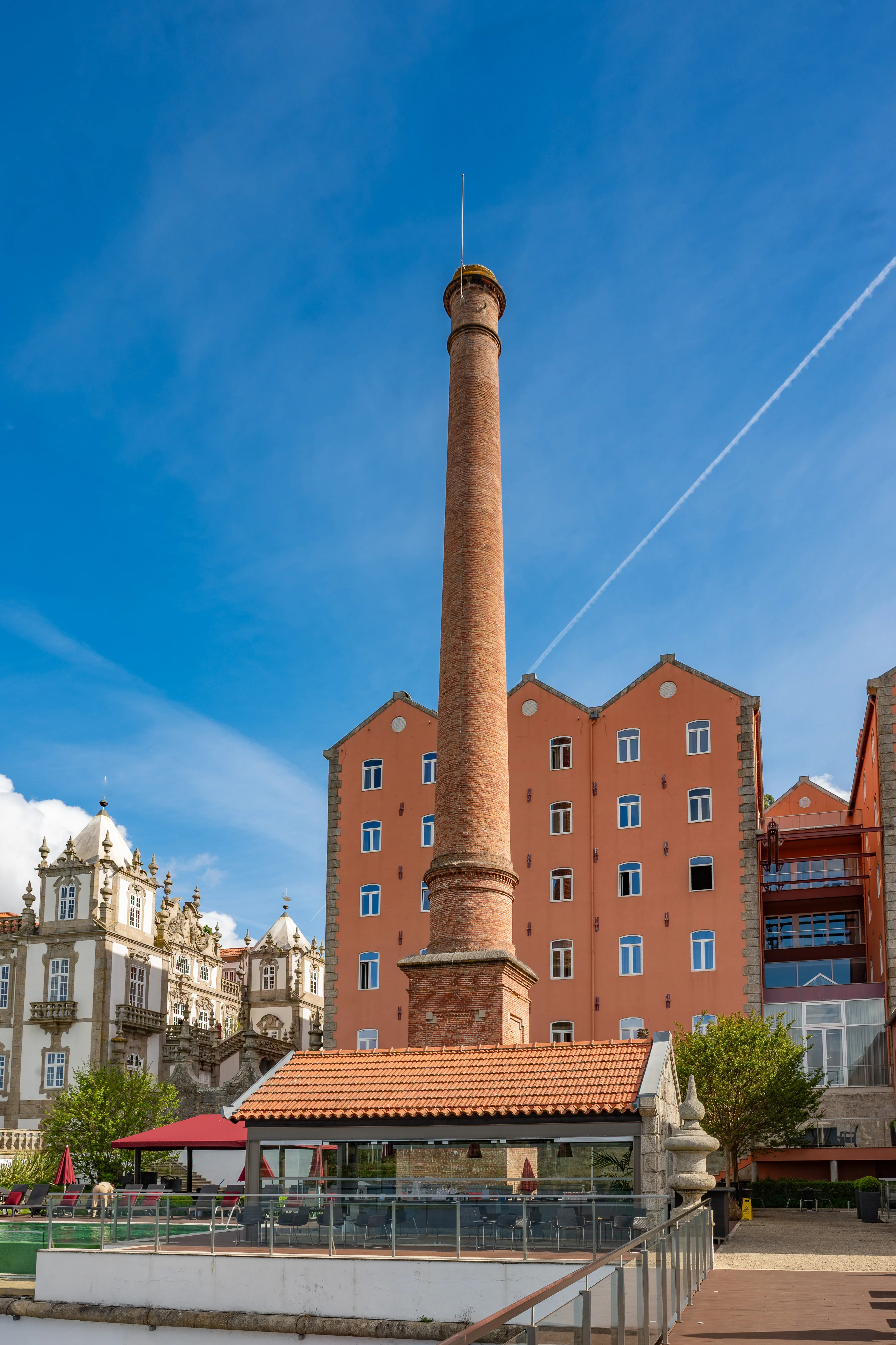 A tall red brick chimney dominates the frame, with a modern orange warehouse building to its right and a decorative white historic building to its left, under a bright blue sky. In the foreground, an outdoor cafe area with tables and chairs is visible.