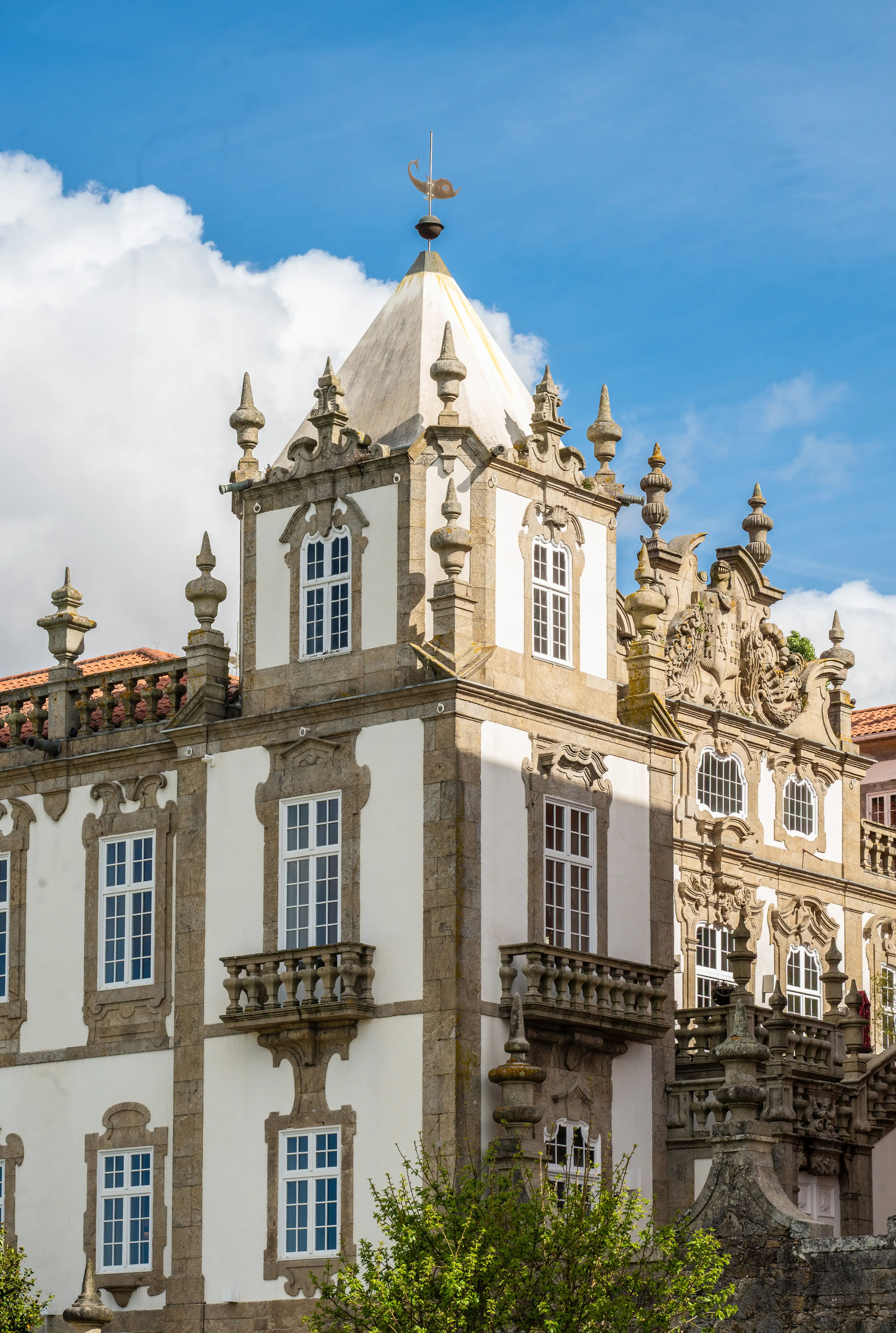 A detailed view of a white and stone building with ornate architectural features, including multiple windows, decorative balconies, pointed pinnacles, and a golden bird weather vane on a pyramidal roof, set against a blue sky with clouds.