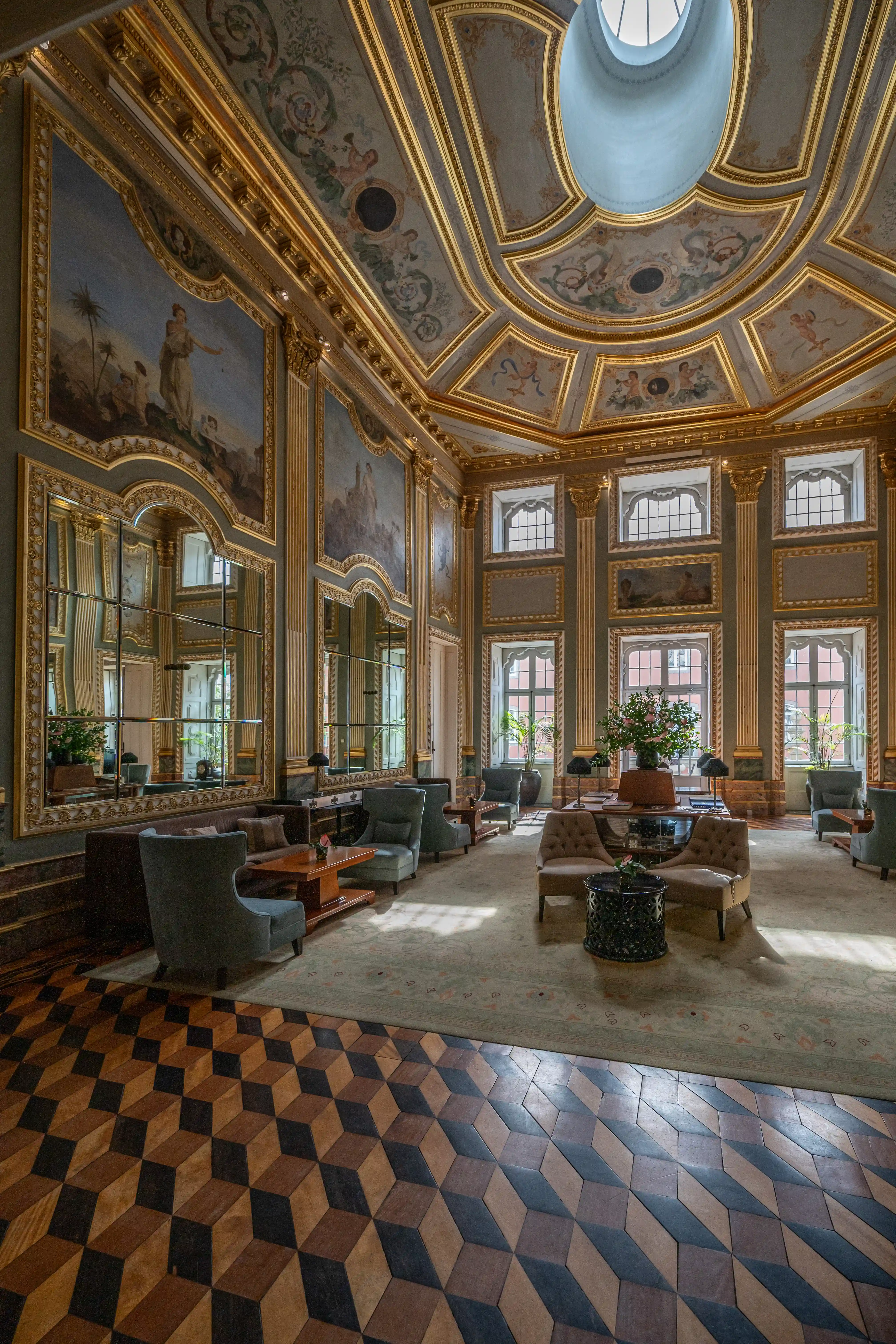 A wide shot of a luxurious drawing room with ornate gilded mouldings, ceiling frescoes, large mirrors, comfortable seating, and a distinctive geometric parquet floor.