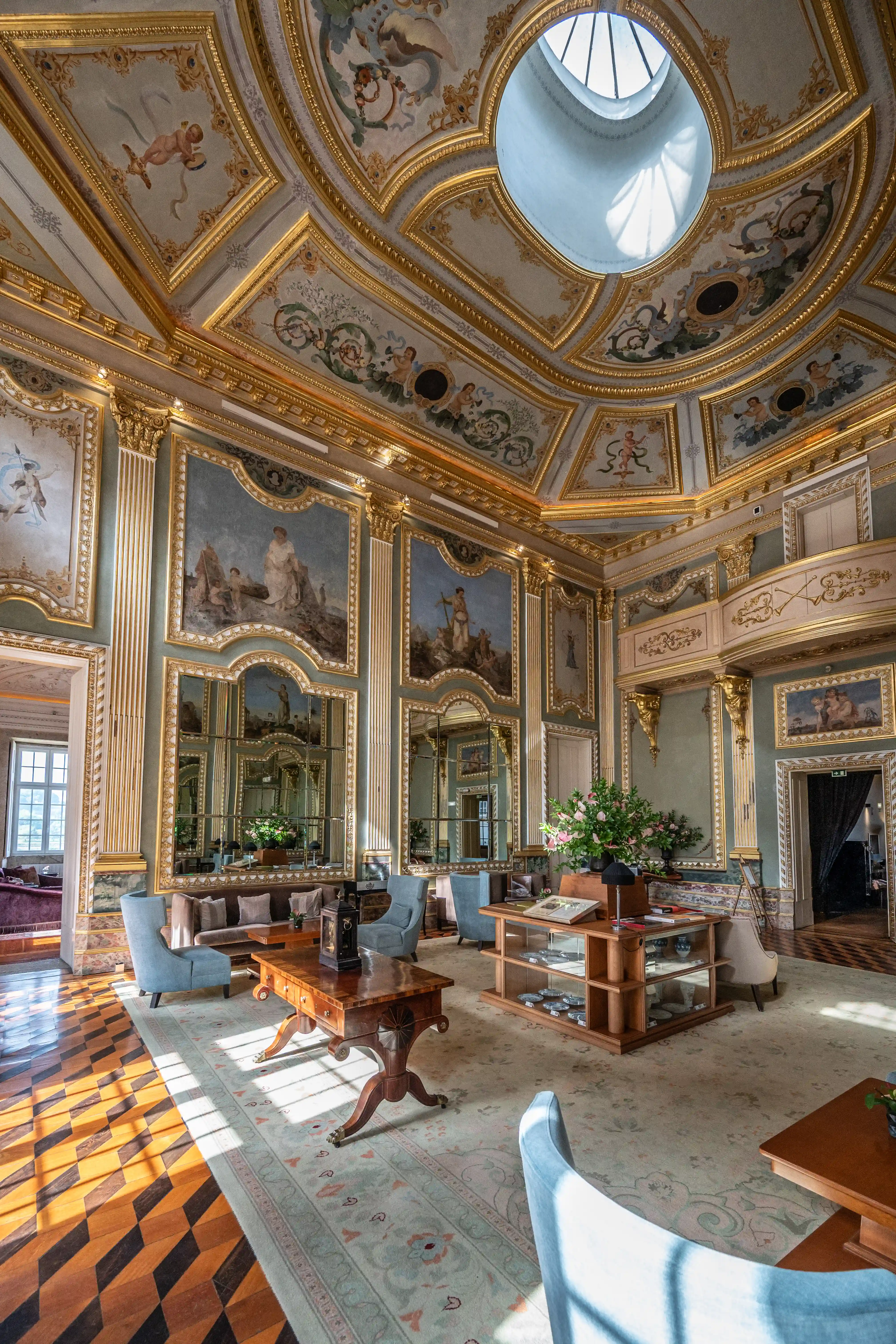 A richly decorated salon with a frescoed domed ceiling featuring gilded mouldings and mythological scenes. Ornate paintings and large mirrors adorn the pale green walls, complemented by fluted columns. Blue armchairs and a sofa surround a dark wooden coffee table. A patterned parquet floor transitions to a patterned carpet. Sunlight streams in from a large skylight and a doorway, illuminating the luxurious interior.