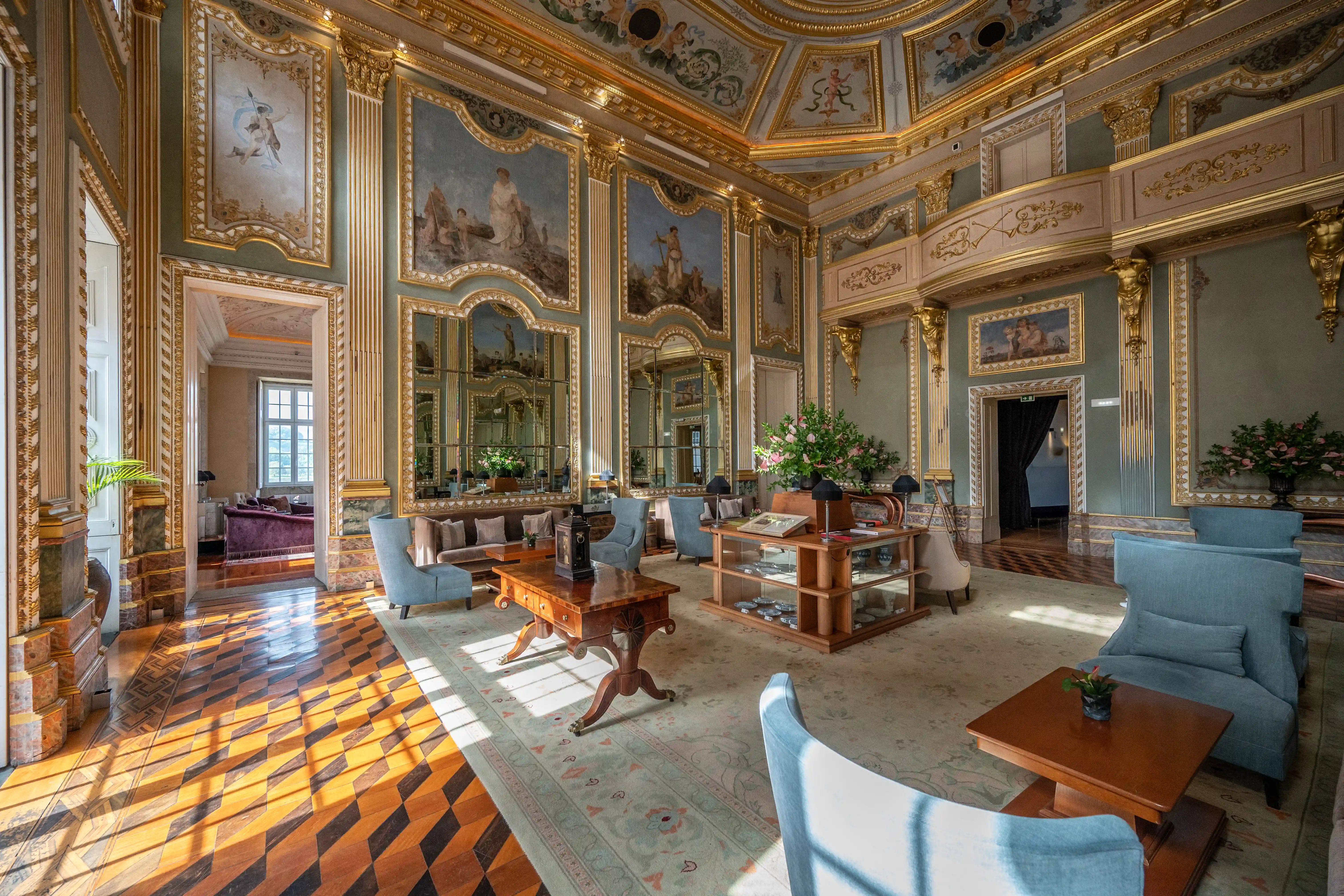 A grand, ornately decorated drawing room with pale green walls, gilded mouldings, and framed frescoes. Several light blue armchairs and a sofa are arranged around wooden tables on a patterned rug. A patterned parquet floor is visible in the foreground, with sunlight casting shadows. Mirrors and floral arrangements add to the luxurious atmosphere.