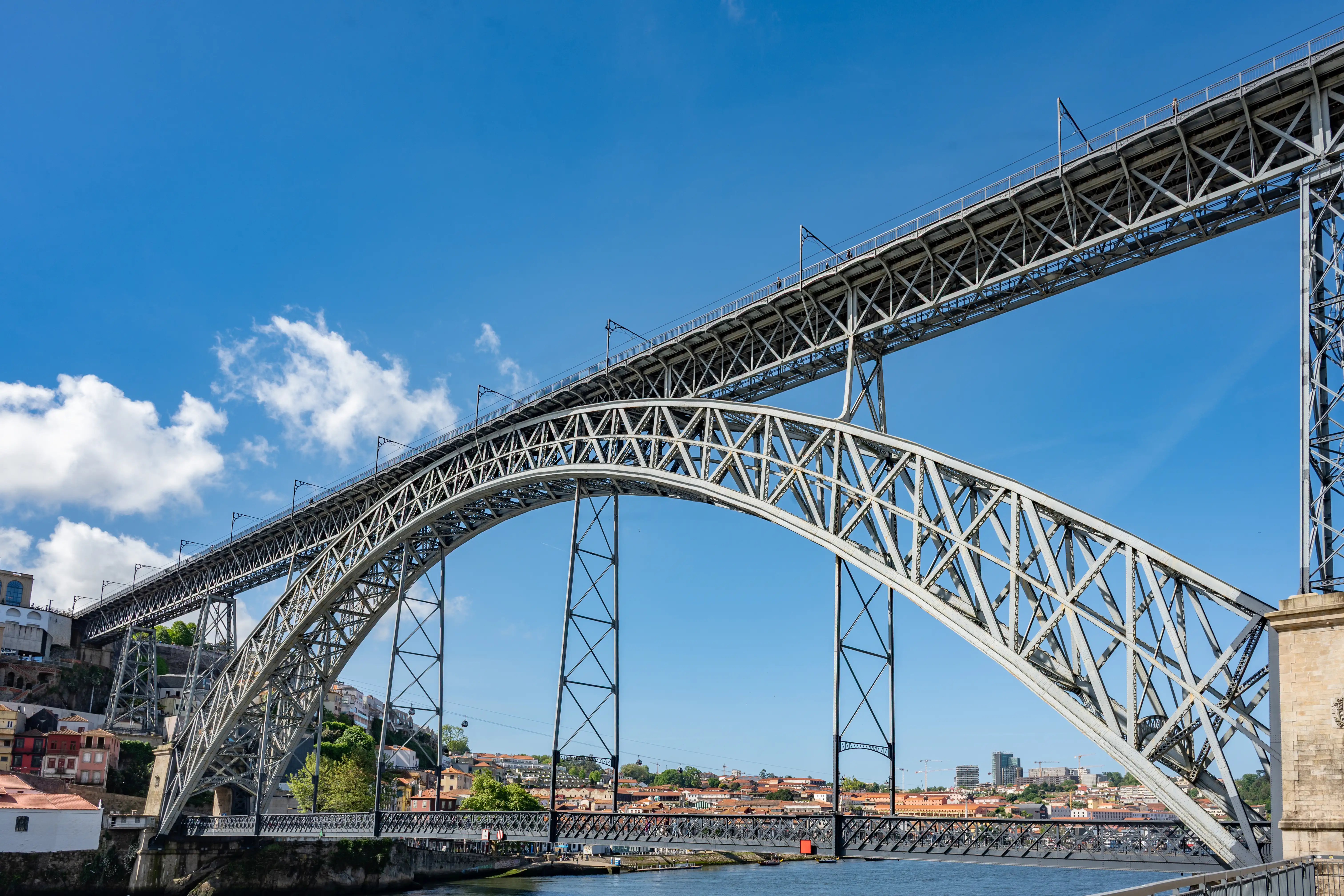 A wide shot of the Dom Luís I Bridge in Porto, Portugal, showcasing its impressive twin metal arches against a bright blue sky with scattered white clouds. The bridge carries road traffic on its upper level and pedestrians and light rail on its lower level. The historic Ribeira district with its terracotta roofs lines the riverbank on the far side.