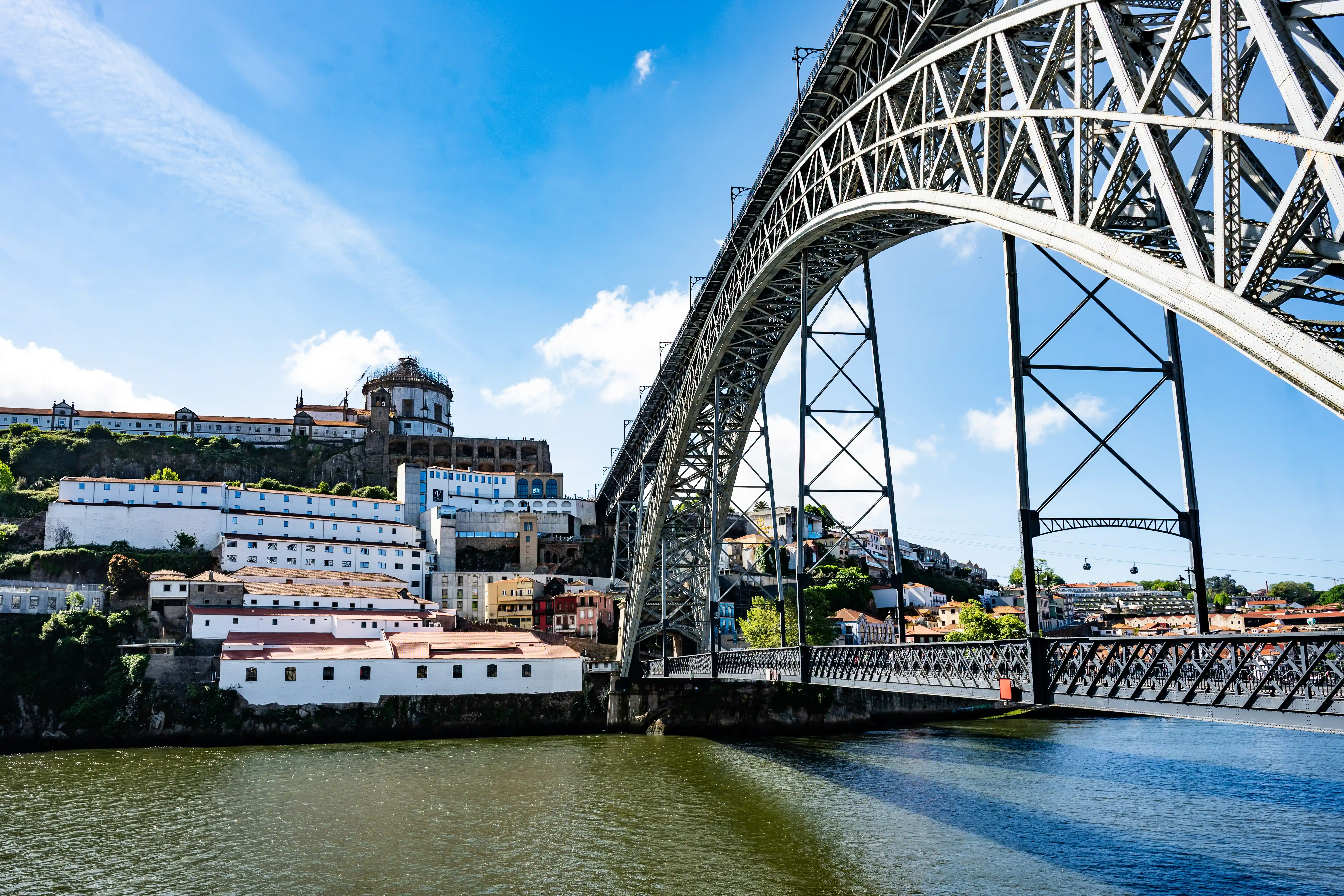 A wide shot of the Dom Luís I Bridge in Porto, Portugal, with its impressive metal arches stretching across the Douro River. The historic buildings of Porto, including the Monastery of Serra do Pilar, are visible on the hillside to the left, bathed in sunlight.