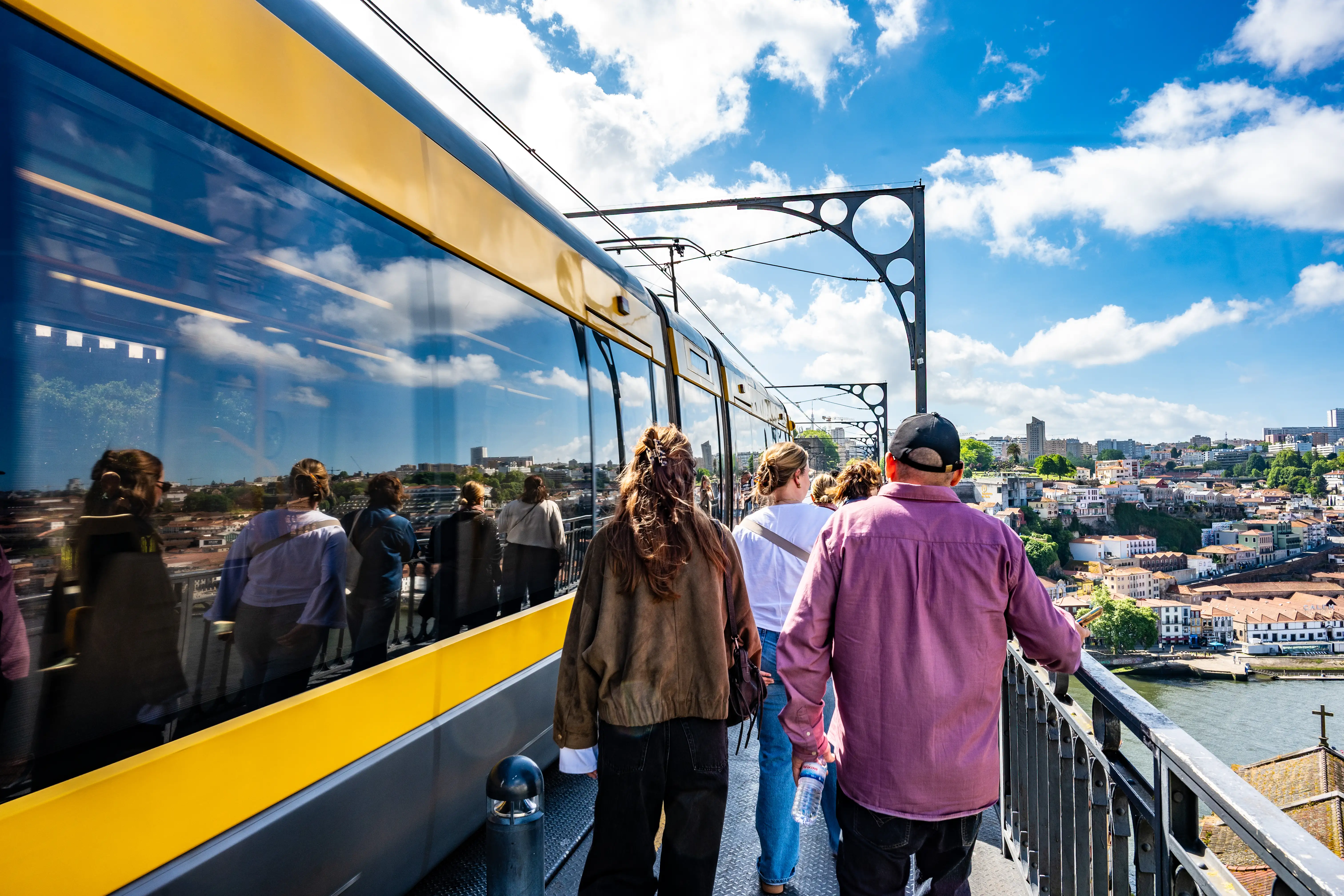 People walking on a bridge next to a yellow tram, with a view of the cityscape of Porto, Portugal, in the background under a blue sky with white clouds.