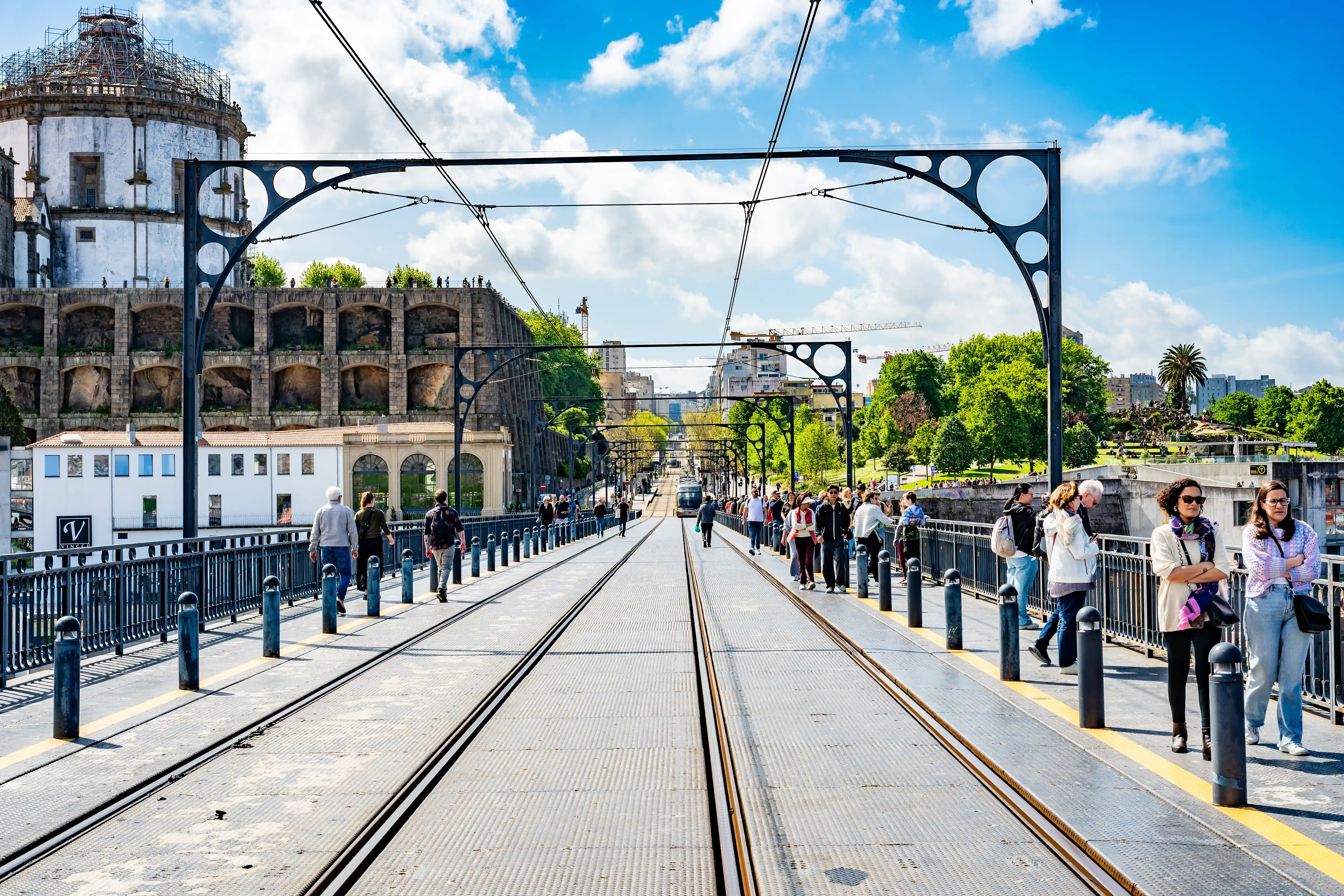 A wide shot of the Dom Luís I bridge in Porto on a sunny day, with a tram in the distance and many people walking across the upper deck. The historic buildings and lush greenery of the city are visible in the background under a blue sky with white clouds.