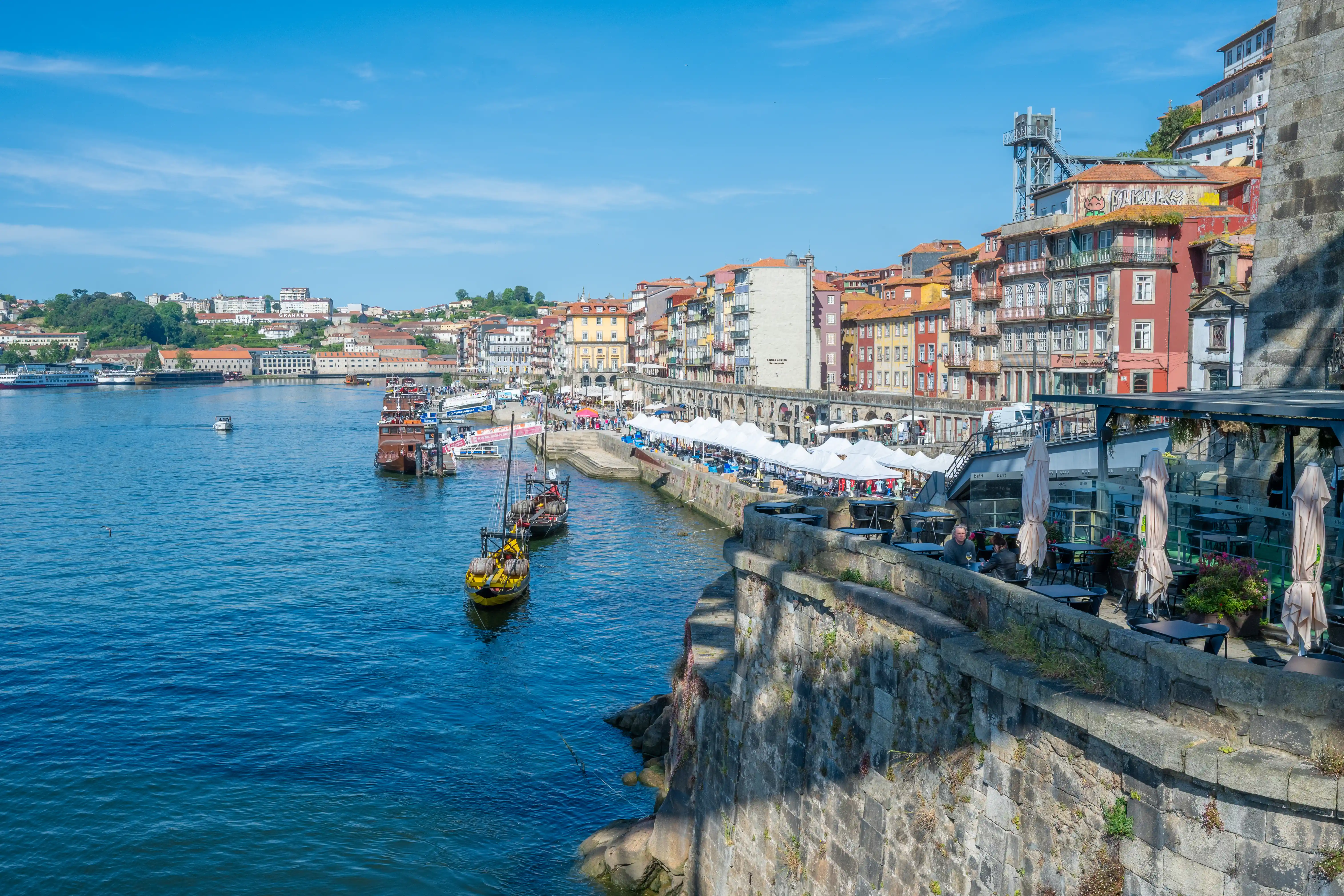 A wide shot of the colourful buildings along the Douro River in Porto, Portugal. Traditional wooden boats, including a yellow Rabelo boat, are docked in the blue water. An outdoor cafe with tables and umbrellas sits on a stone wall overlooking the river.