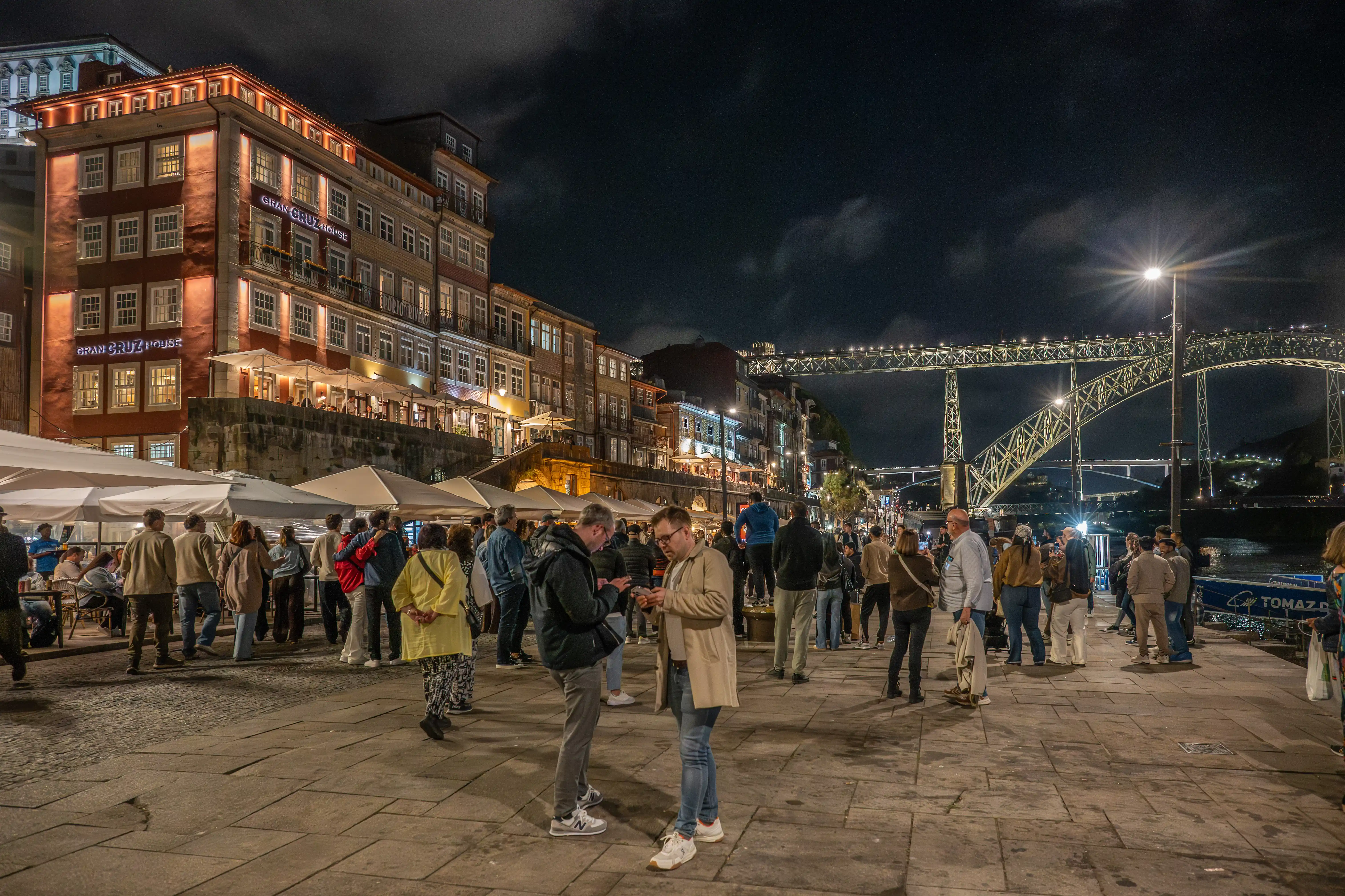 A wide shot at night of the Ribeira district in Porto, Portugal. People are gathered on a cobblestone promenade in front of illuminated historic buildings. The Dom Luís I Bridge is visible in the background, spanning the Douro River.