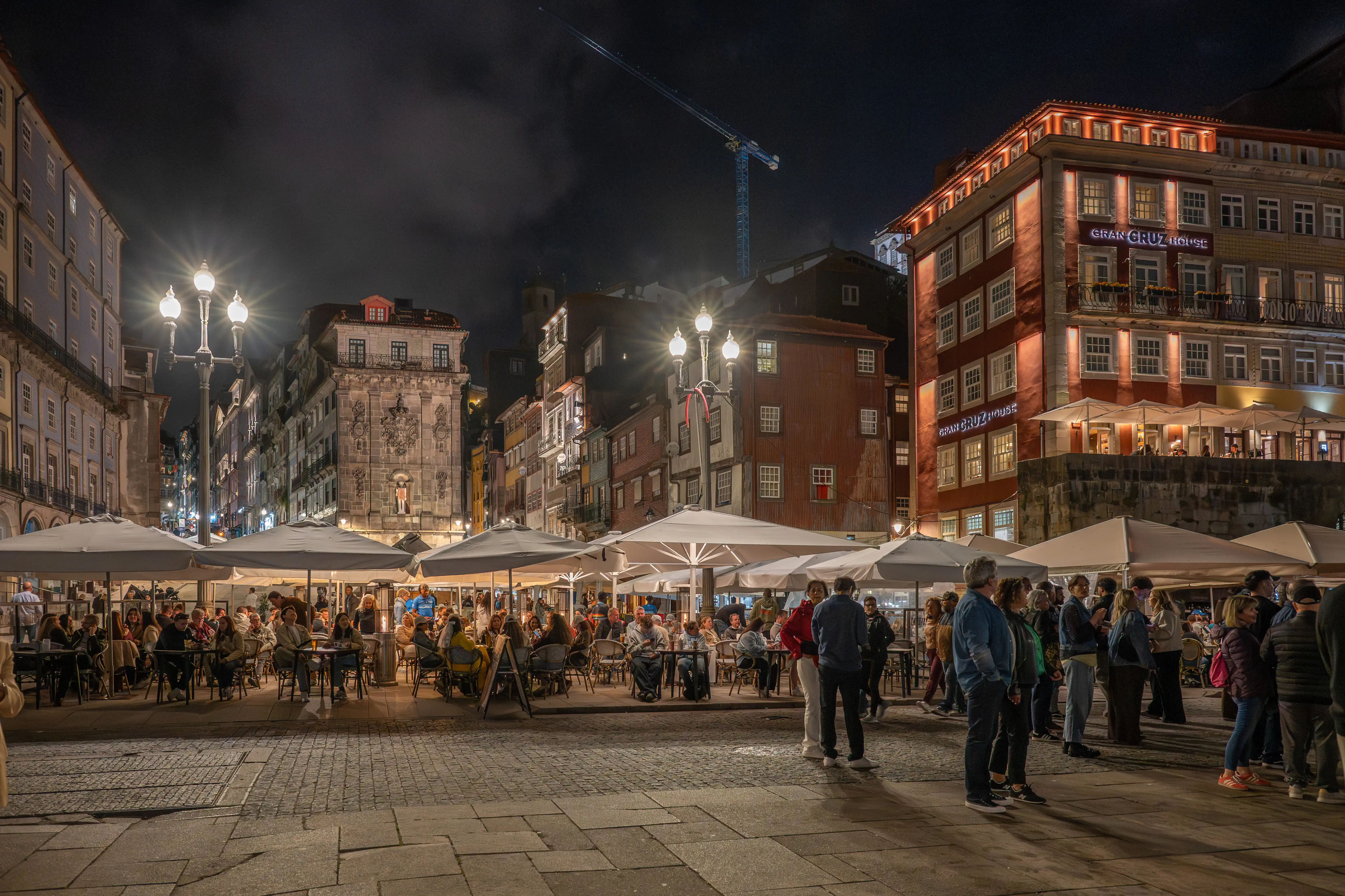  A lively European square at night, with people dining outdoors under umbrellas. Buildings with traditional architecture are lit up, and a construction crane is visible in the distance.