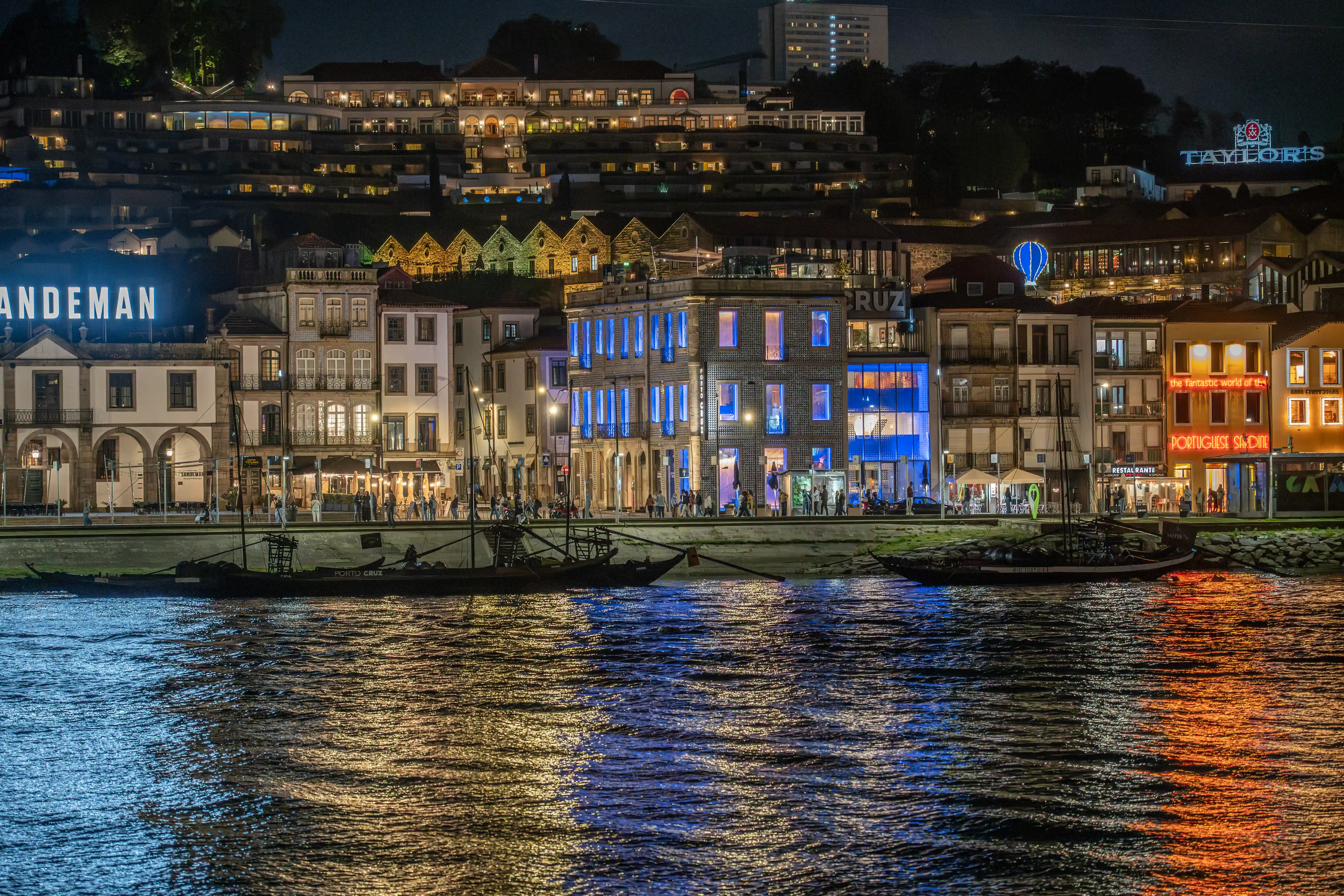 A nighttime shot of Porto's waterfront. Buildings along the river are lit up with bright lights, including prominent signs for Sandeman and Taylor's port wine lodges. Traditional wooden Rabelo boats are docked on the dark, rippling water of the Douro River, reflecting the colourful lights from the buildings and the sky.
