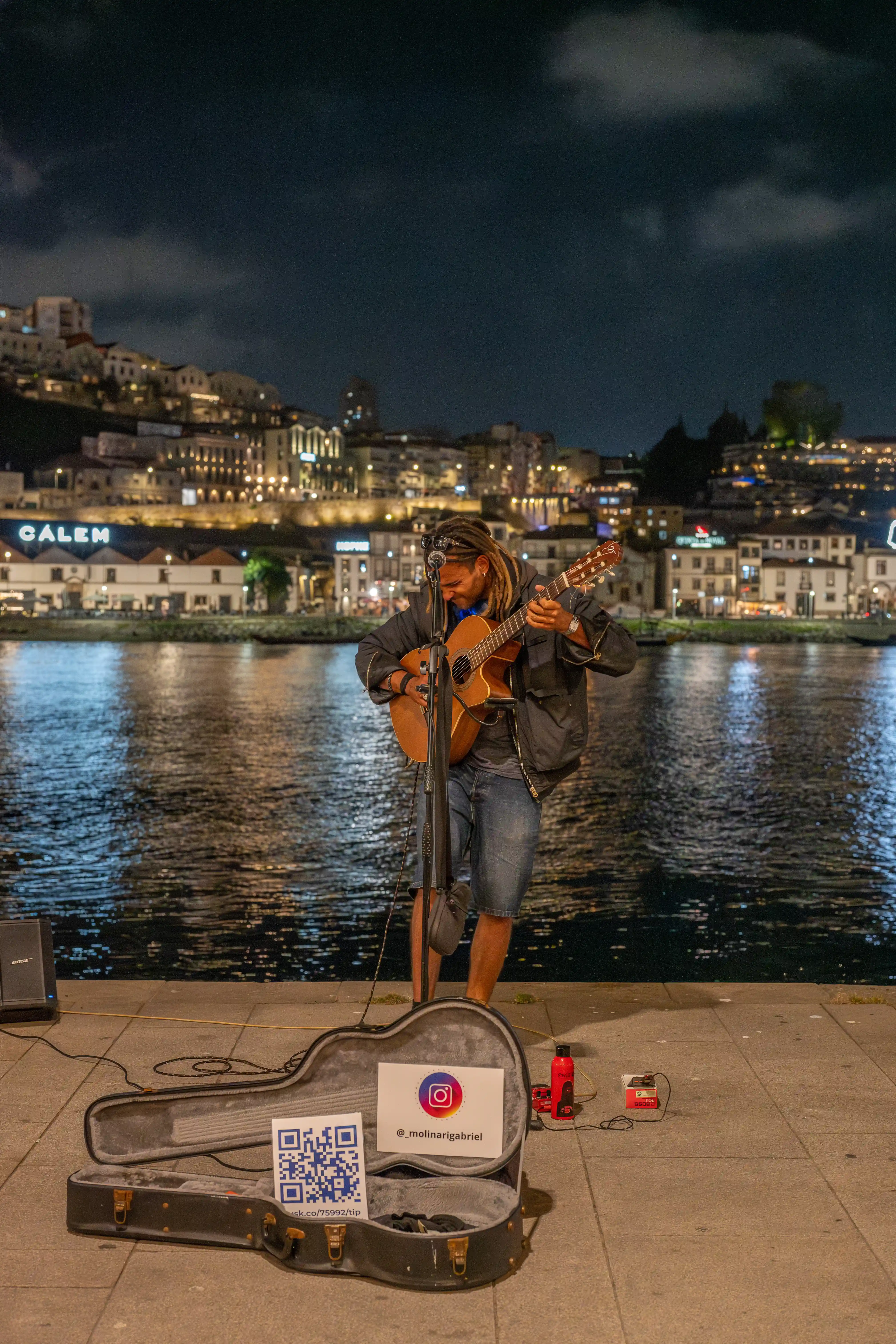 A street musician plays an acoustic guitar by a river at night, with a cityscape in the background and an open guitar case with a QR code and Instagram handle in the foreground.