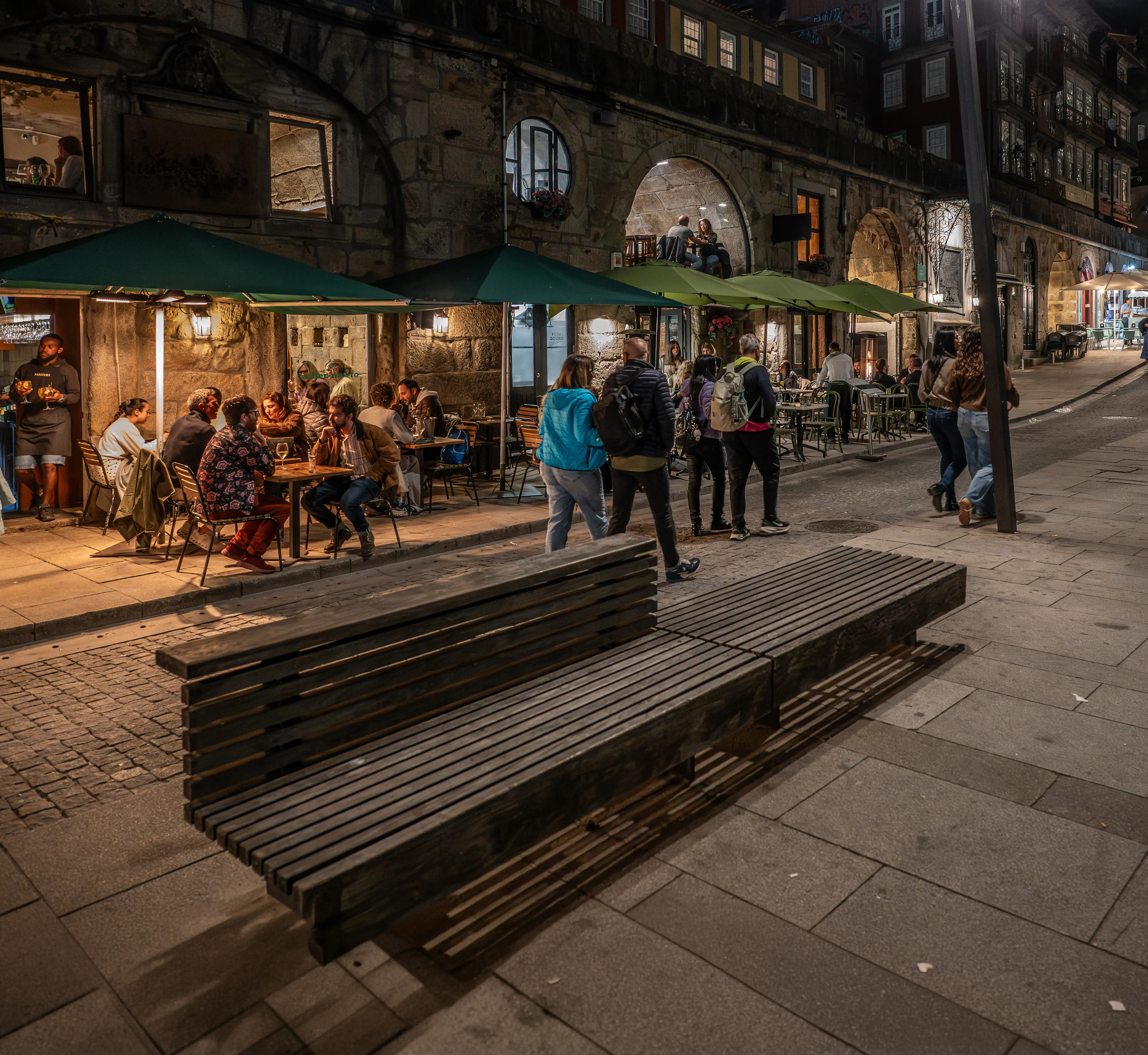 A group of people dining at tables outside a restaurant at night, with green umbrellas providing shade. The restaurant is housed in a historic stone building with arched windows. People are walking along a cobbled street in the foreground, and a modern wooden bench is visible.