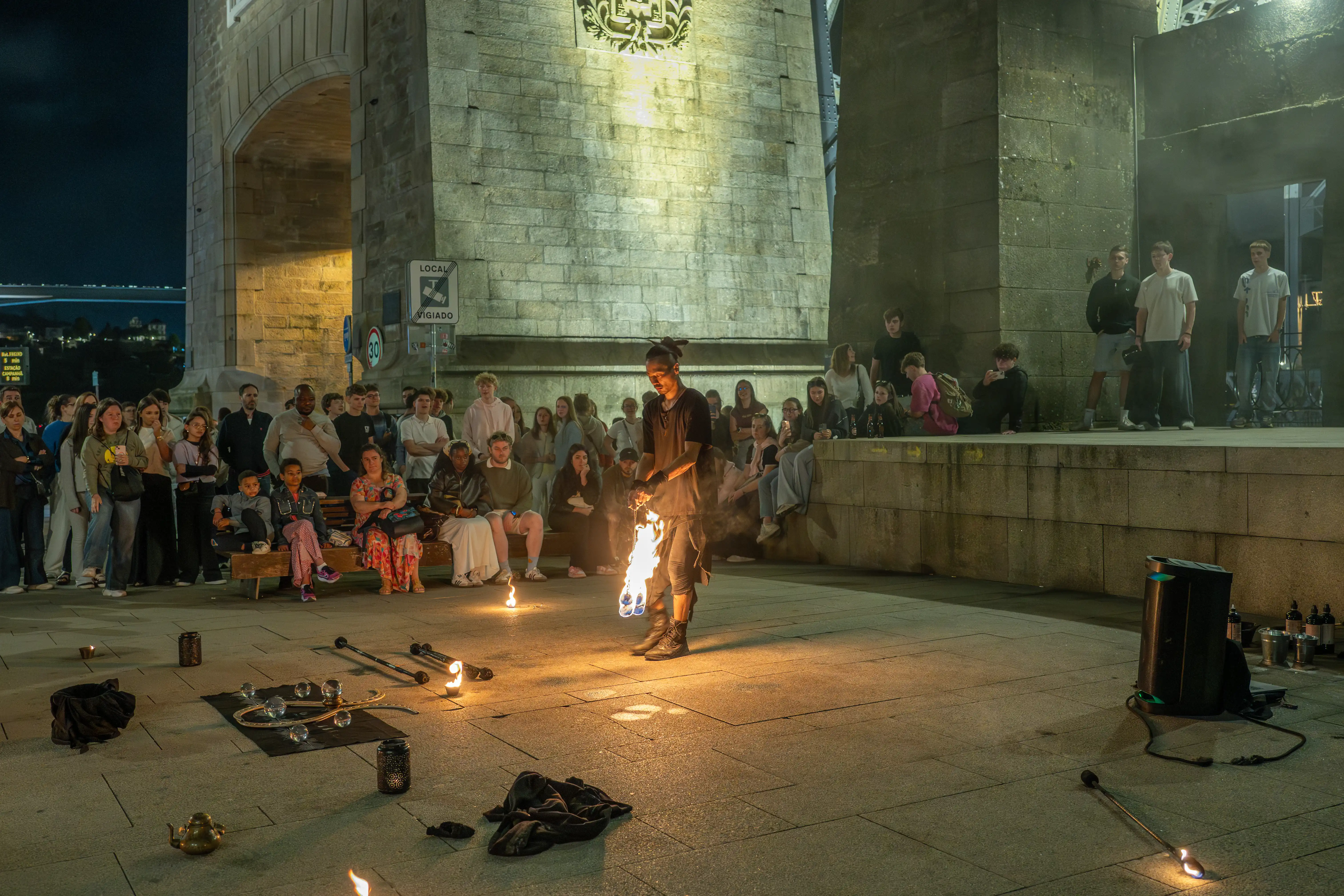 A fire performer with dreadlocks, wearing dark clothing, juggles flaming props in front of an audience seated on a raised stone wall and standing on the ground, under a large stone bridge structure at night. Flames from the props cast a warm glow on the performer and the surrounding pavement, with several other props and performance equipment visible on the ground.