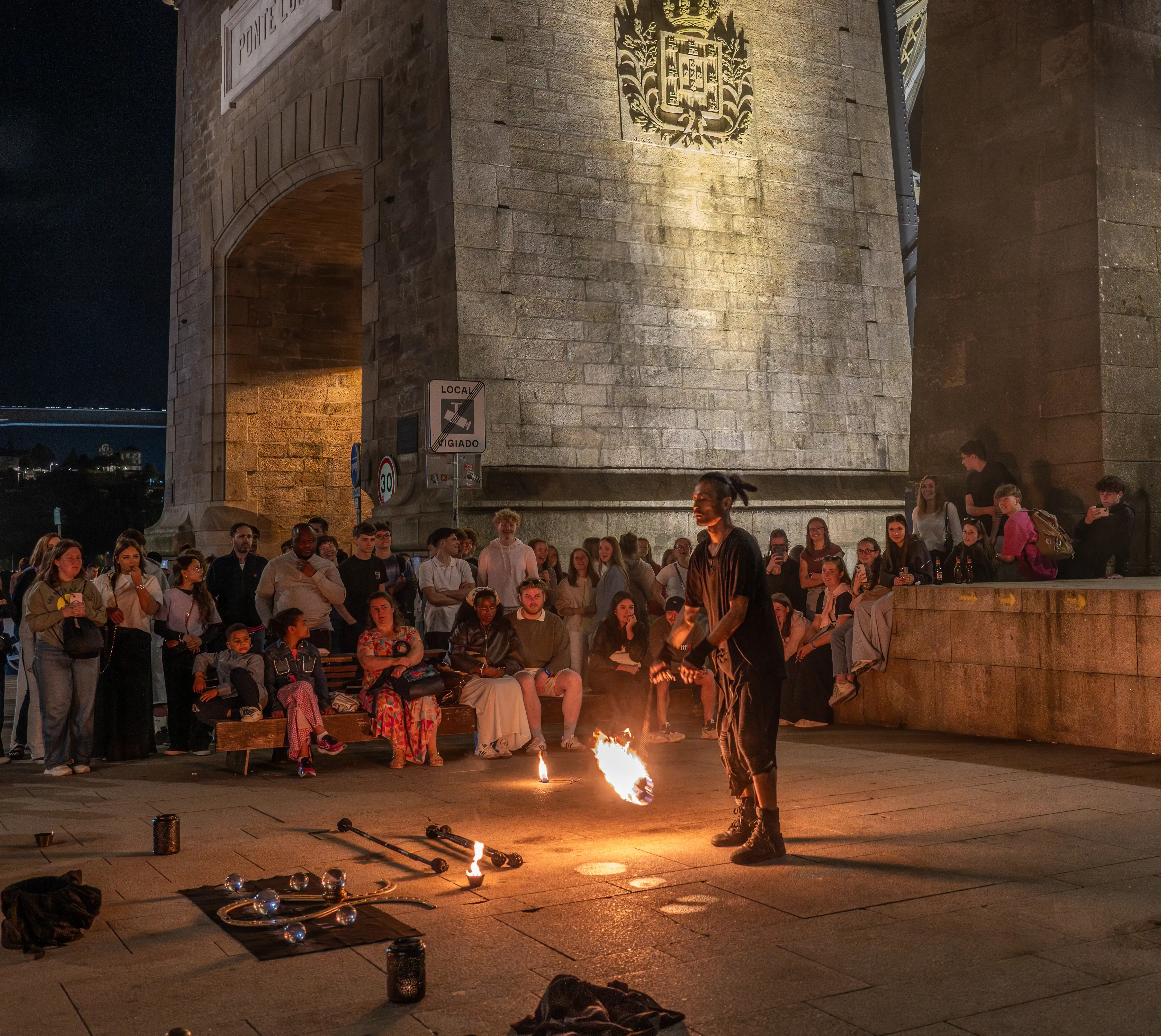 A street performer entertains a crowd at night under stone arches, juggling with a flaming prop. Spectators of all ages watch, some sitting on benches, illuminated by the fire's glow. Crystal balls and juggling sticks are visible on the ground.