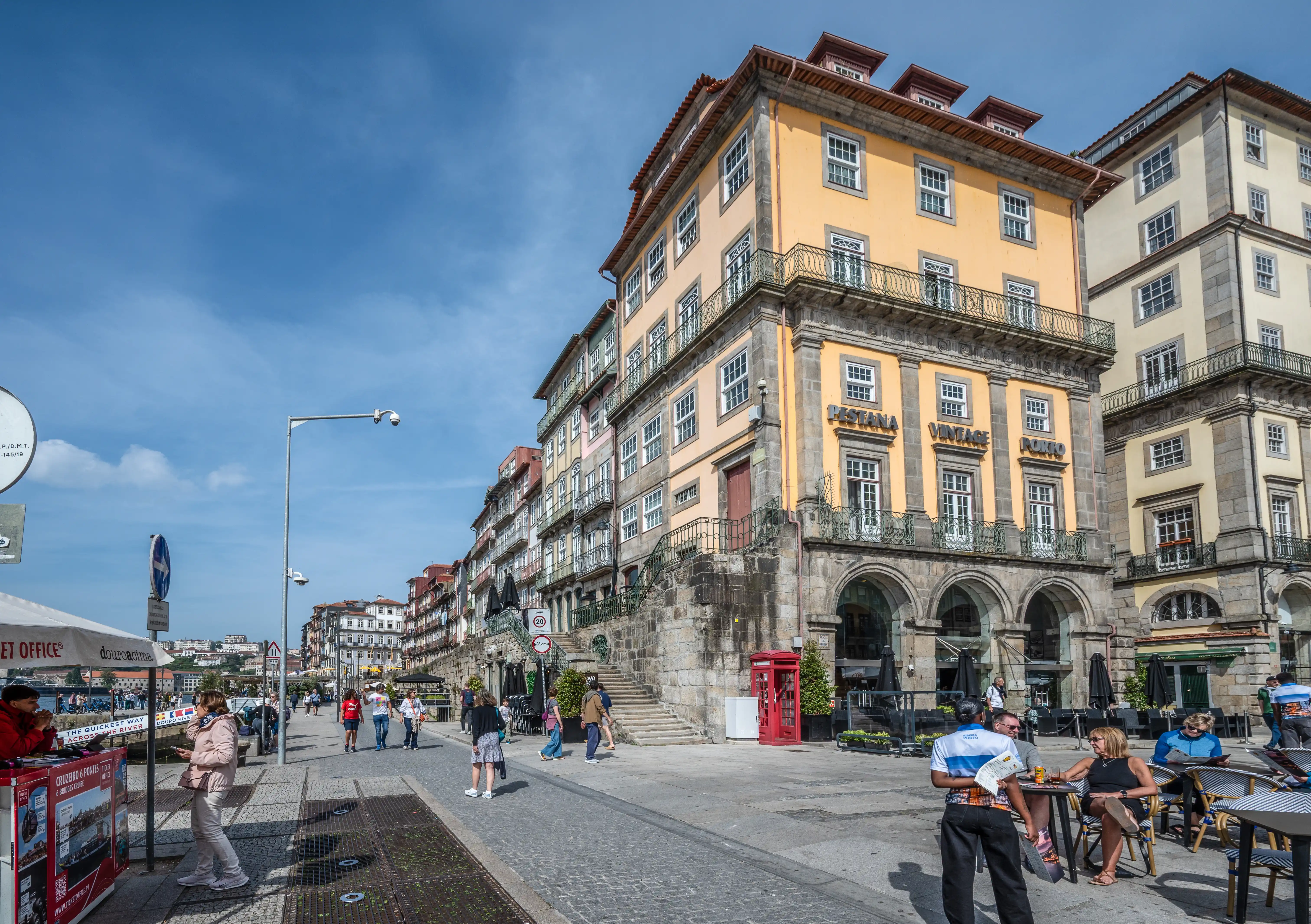 A sunny day on the Douro River waterfront in Porto, Portugal, featuring colourful historic buildings, a cobblestone promenade with people, outdoor cafe seating, a red telephone box, and a tour boat ticket booth.