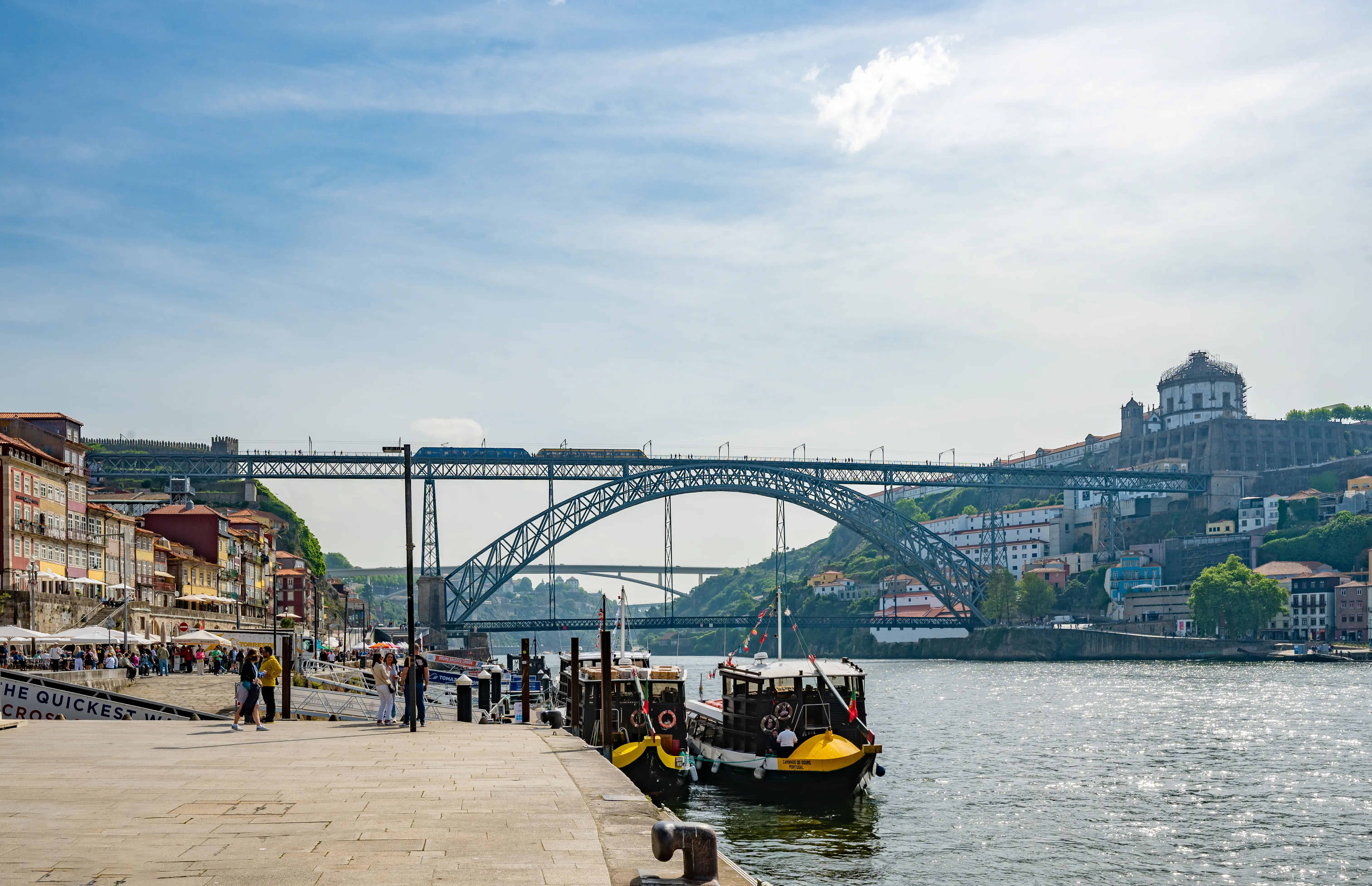 A wide shot of the Dom Luís I Bridge in Porto, Portugal, with a train crossing the upper deck. Colourful buildings line the Douro River waterfront on the left, and two traditional Rabelo boats are moored at the quay. The Serra do Pilar Monastery is visible on the hill in the background under a blue sky with clouds.