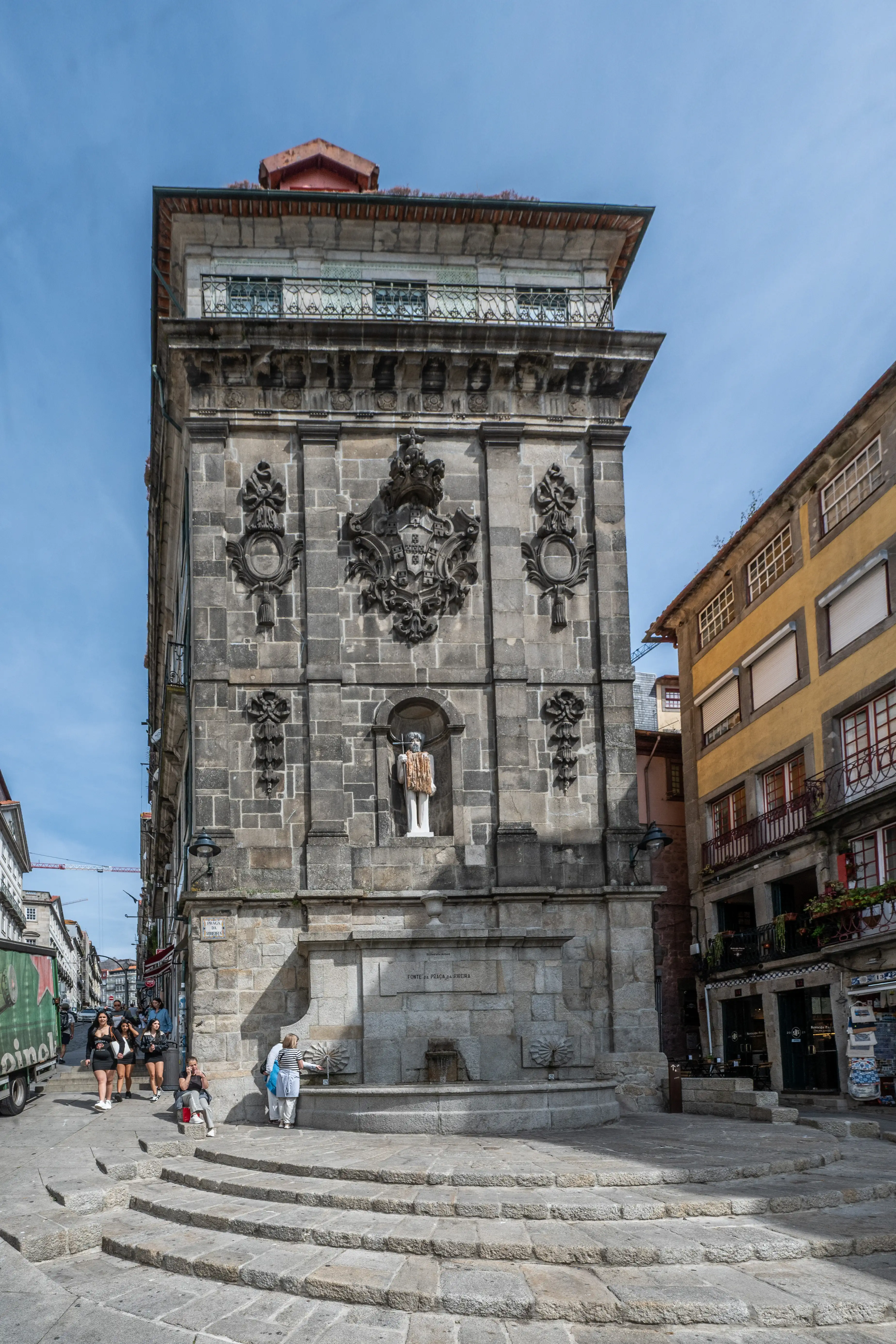 A stone fountain, the Fonte da Praça da Ribeira, is set into a historical building with ornate carvings and a statue in a niche. A wide stone staircase leads up to the fountain, and people are walking nearby on a sunny day.
