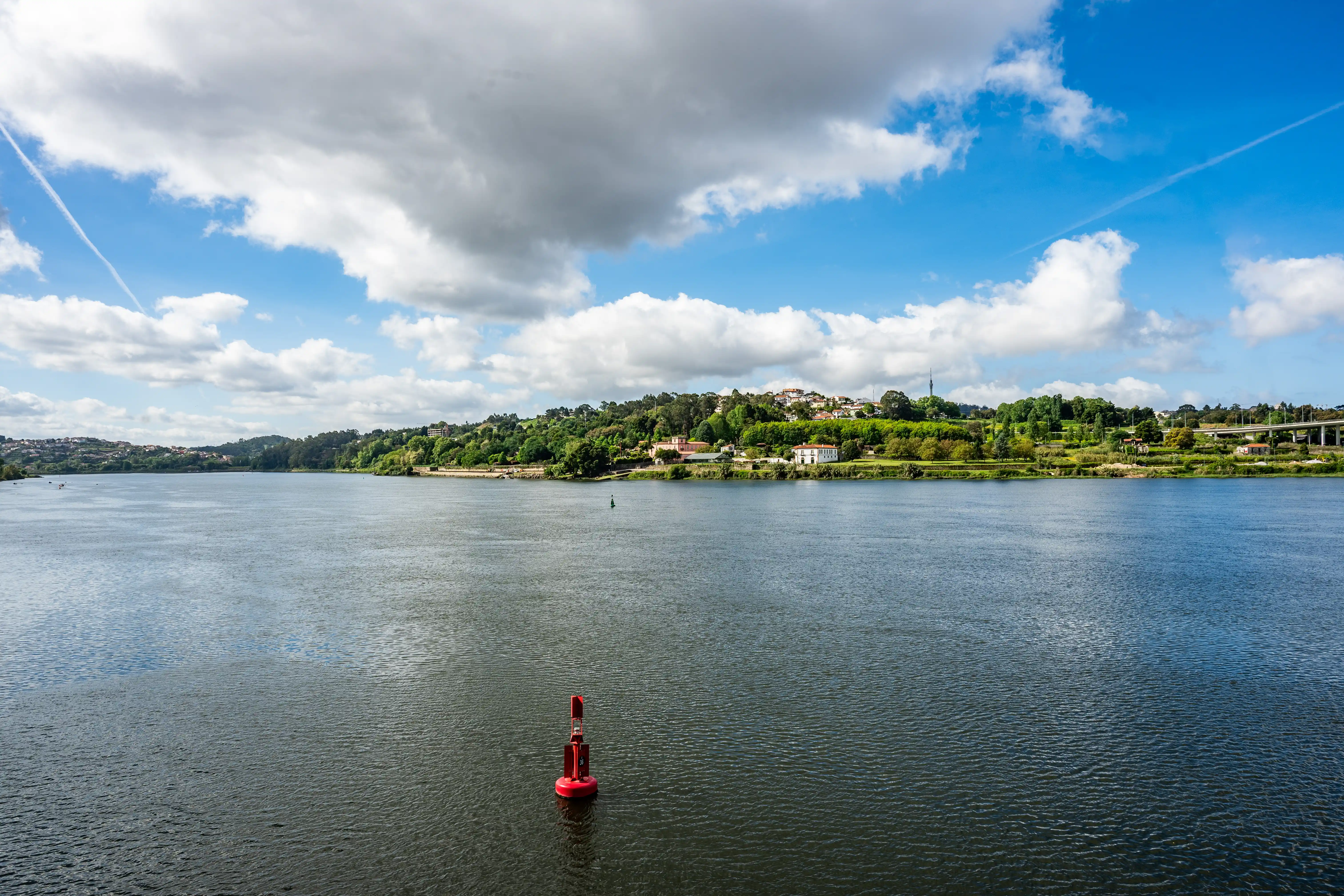 A wide river with a red navigation buoy in the foreground and a tree-lined bank with houses and a bridge in the background, under a partially cloudy blue sky.