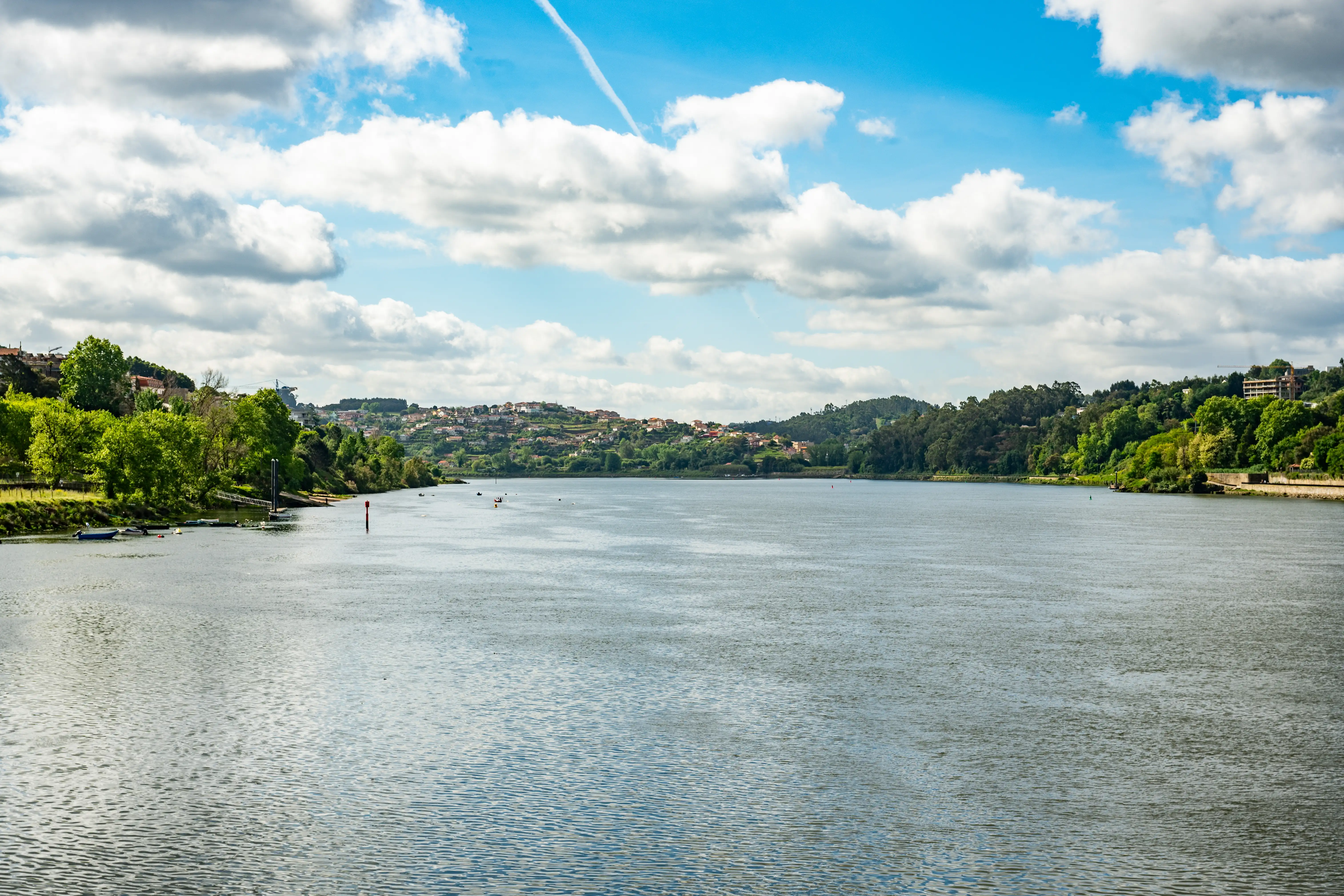 A wide, calm river flows through a landscape of green hills topped with a town of red-roofed houses, under a blue sky with white, fluffy clouds.
