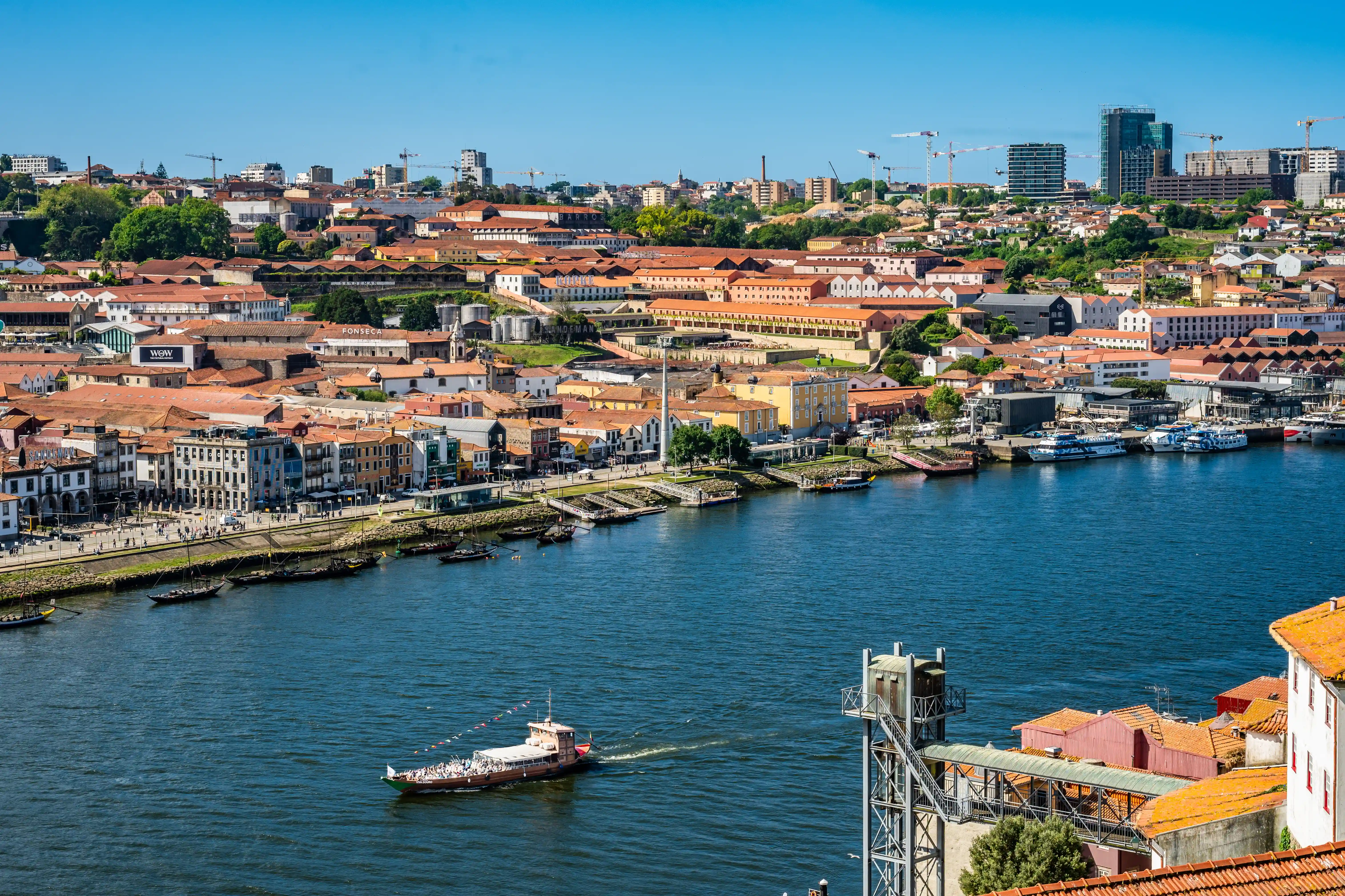  A wide shot of the Douro River in Porto, Portugal, under a bright blue sky. The north bank is lined with buildings featuring red-tiled roofs and colourful facades, with signs for port wine cellars visible. A traditional wooden boat carrying people crosses the river. Several modern boats are docked along the waterfront. In the distance, cranes and newer buildings suggest urban development, contrasting with the historic architecture.