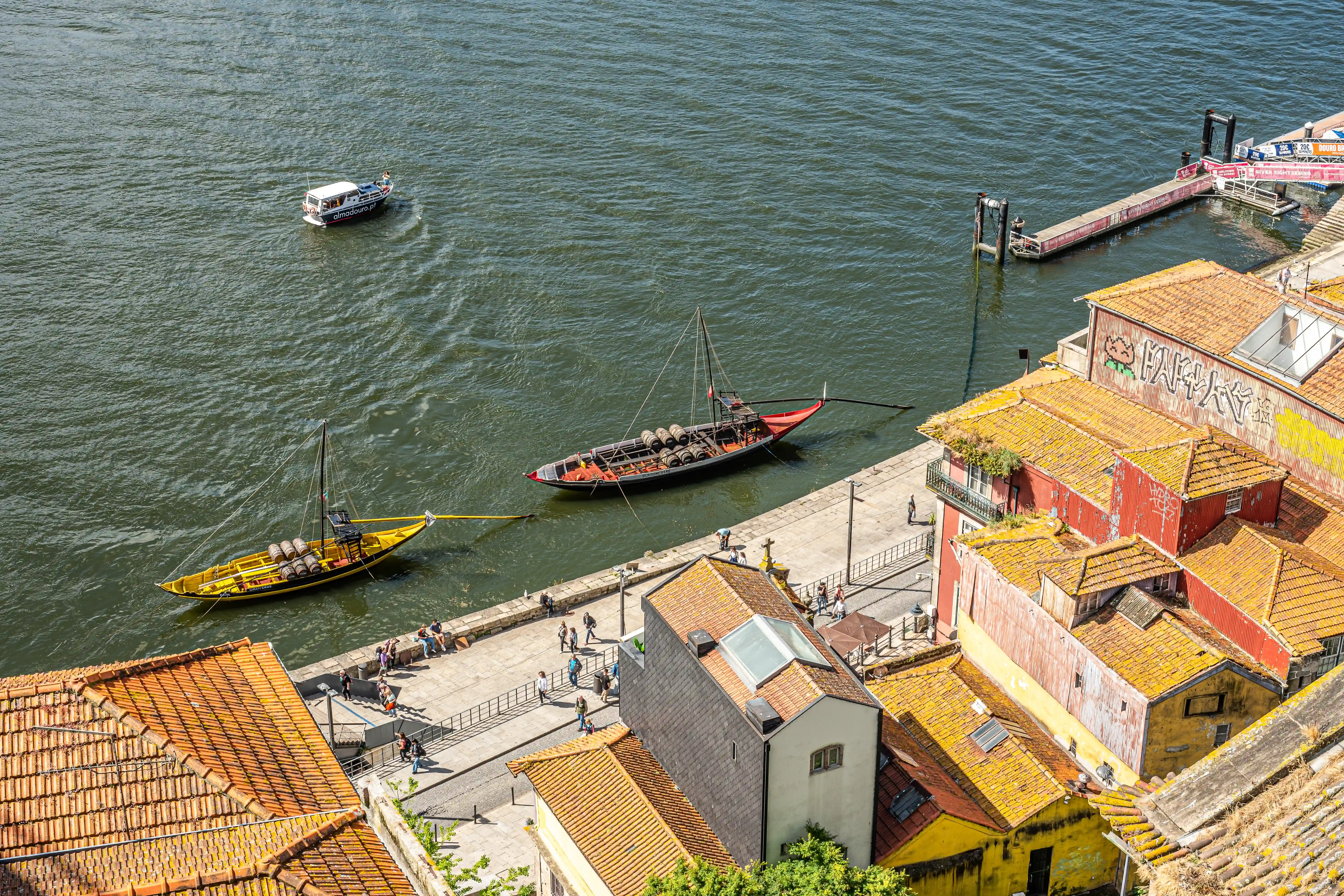 Aerial view of the Douro River in Porto, Portugal, with two traditional Rabelo boats laden with barrels, a white tourist boat, a modern pier, and colourful, historic buildings with tiled roofs lining the riverside promenade where people are walking.
