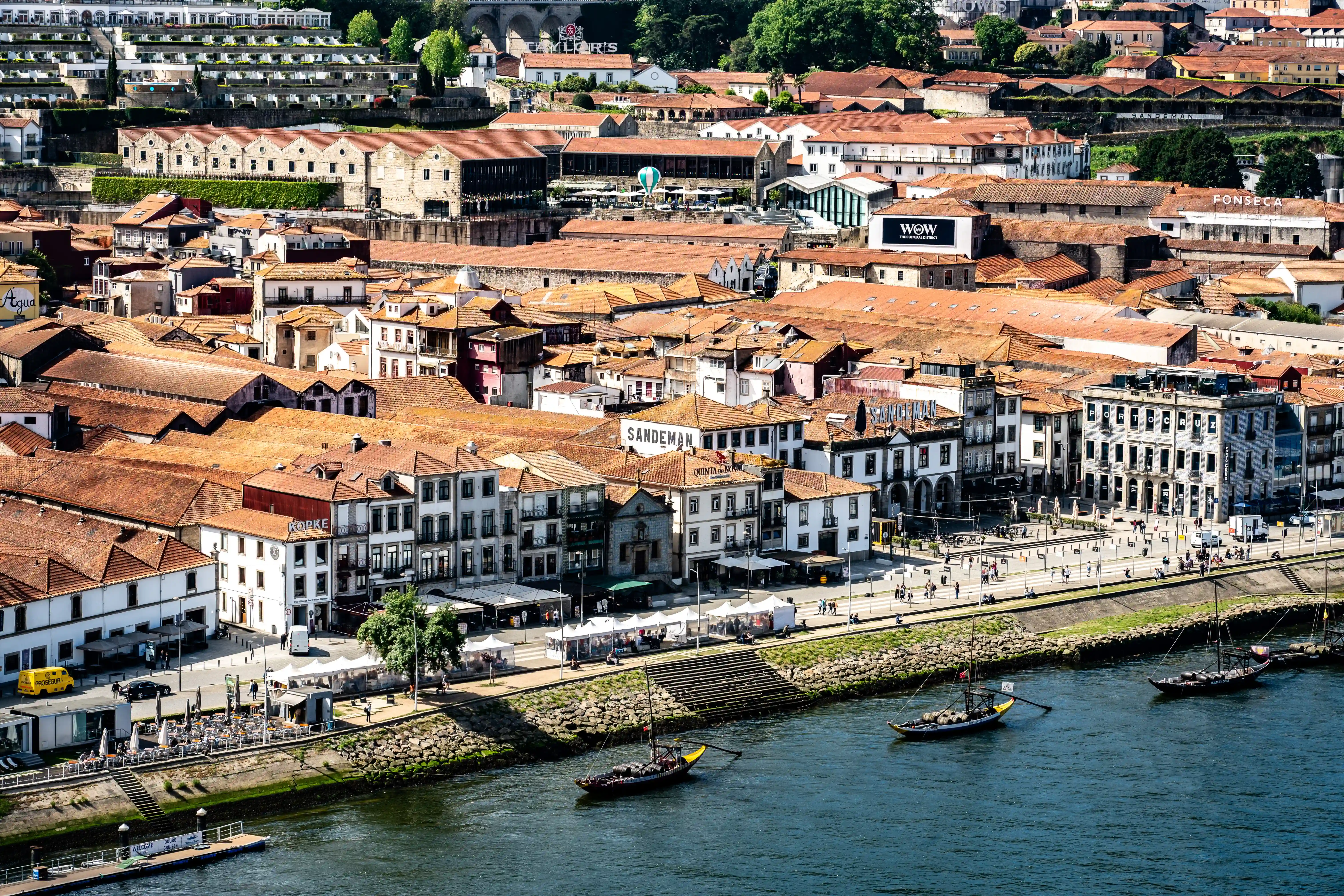 An elevated view of the Gaia district in Porto, Portugal, showing numerous terracotta-roofed buildings, including prominent port wine cellars like Sandeman and Fonseca, lining the Douro River. Traditional Rabelo boats are moored on the river, and a bustling promenade with cafes and people is visible along the waterfront. The background reveals more of the city's architecture and green hills.