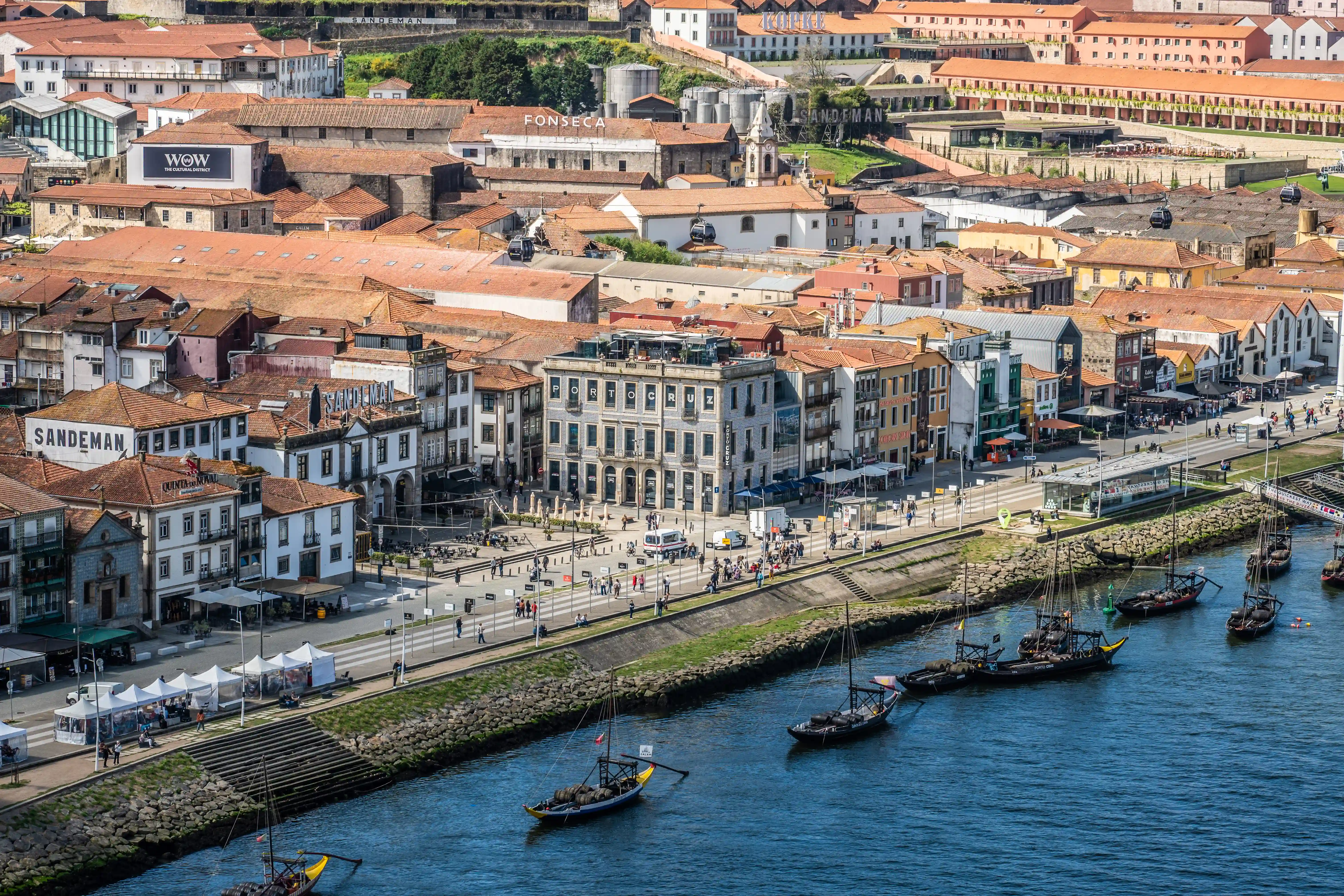 A wide shot of the historic Ribeira district in Porto, Portugal, overlooking the Douro River. Traditional buildings with red tile roofs crowd the waterfront, where a promenade is busy with people. Several Rabelo boats, some carrying barrels, are on the blue water, with cable cars visible in the background.