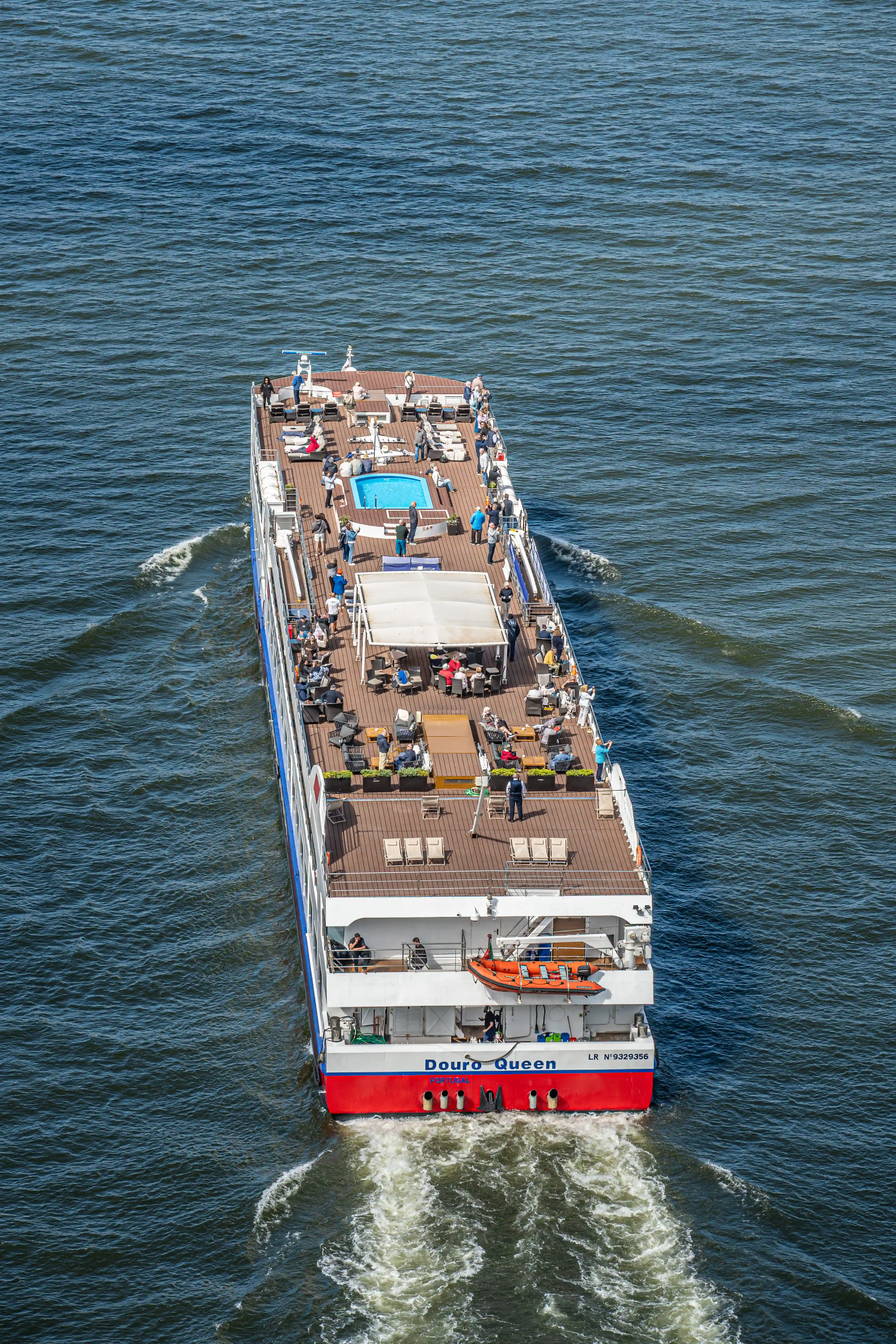 Aerial view of the Douro Queen river cruise ship sailing on the water, showing passengers relaxing on the sundeck near a small swimming pool, with an orange lifeboat visible on the stern.