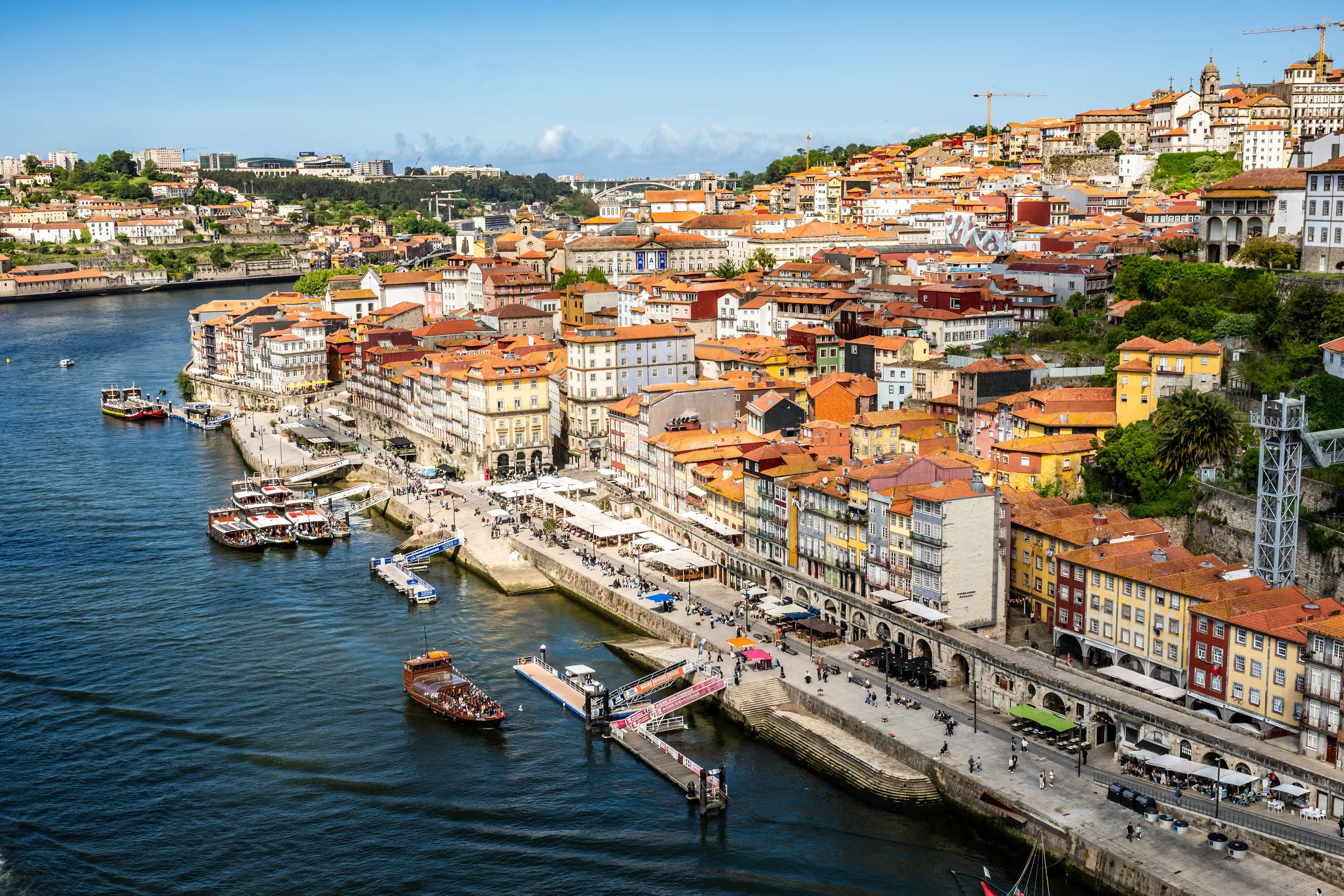 Aerial view of the colourful buildings and terracotta rooftops lining the Douro River in Porto, Portugal, with boats and people visible along the waterfront promenade under a clear blue sky.