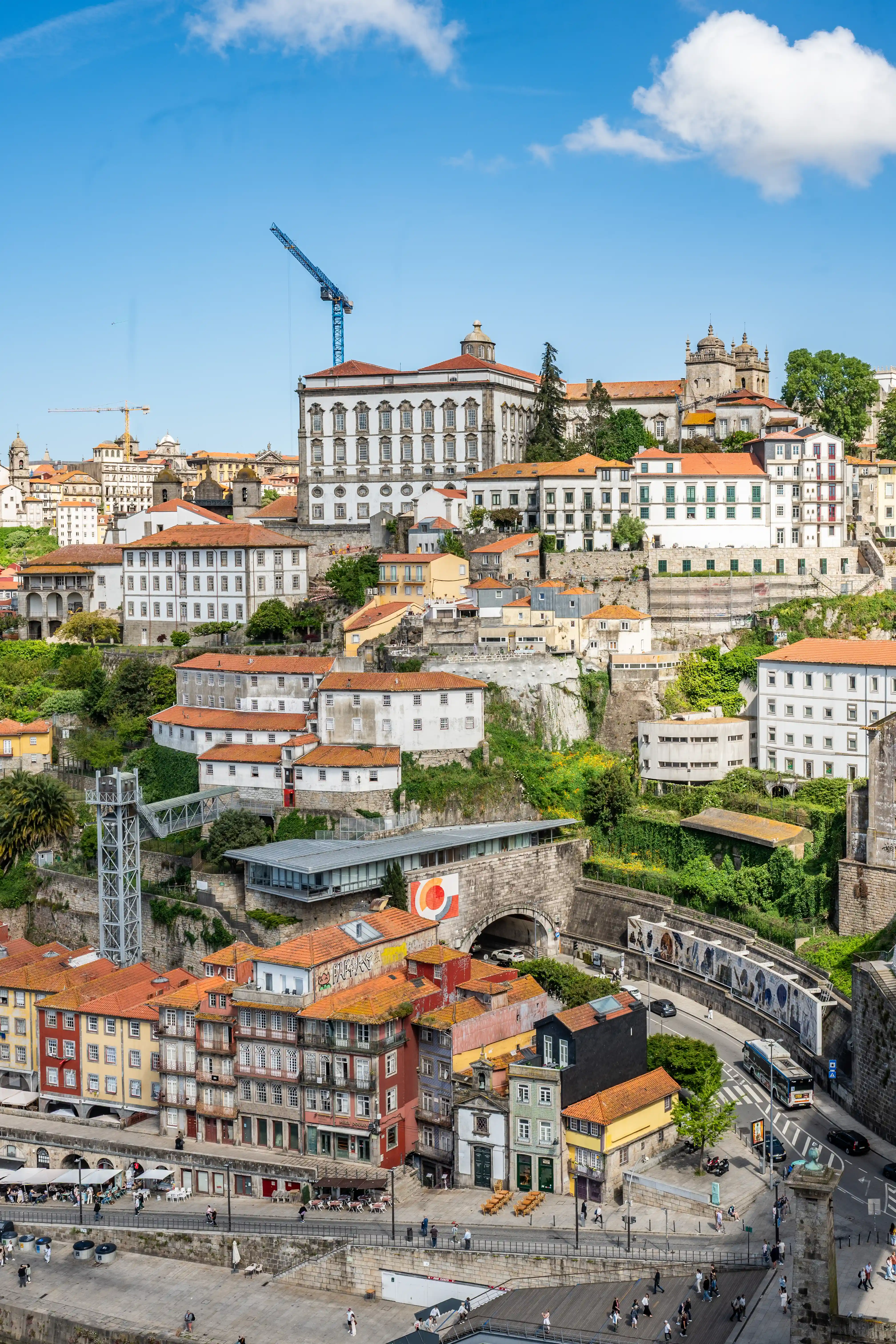 A vibrant cityscape of Porto, Portugal, unfolds under a clear blue sky dotted with fluffy white clouds. The image captures the city's distinctive riverside architecture, a charming mix of colourful, terracotta-roofed buildings cascading down hillsides. Prominent construction cranes, one a striking blue, suggest ongoing development amidst the historic setting. The Douro River is visible at the bottom, with a promenade lined with people and outdoor seating areas. Cobblestone streets and a bridge with a tunnel add depth and character to the scene.