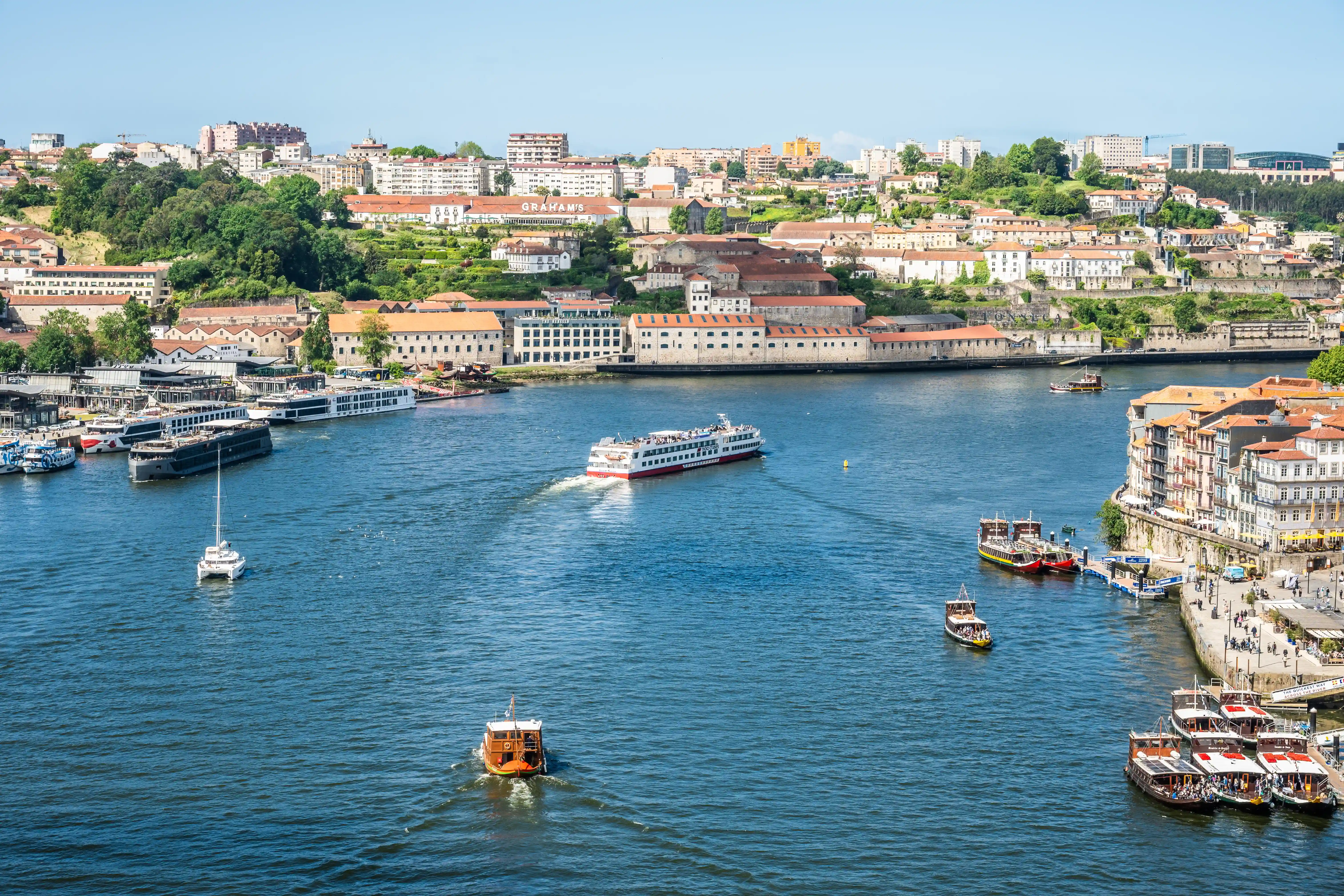 A wide shot overlooking the Douro River in Porto, Portugal, on a sunny day. Several boats, including large passenger ferries and smaller tour boats, are visible on the water. The city's distinctive buildings with red-tiled roofs and white facades rise up from the riverbank, set against a backdrop of green hills.