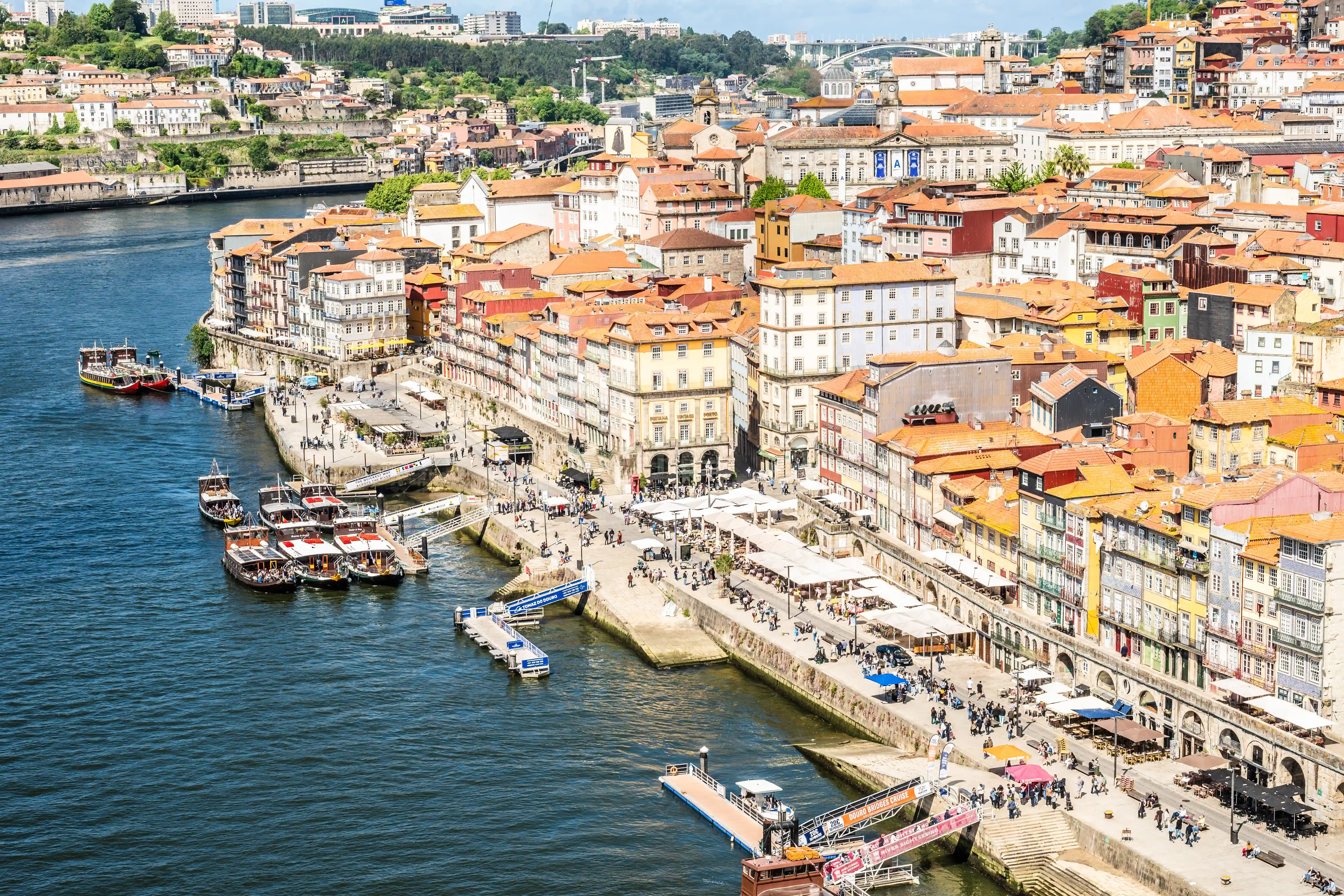 A high-angle view of the colourful, densely packed buildings along the Douro River in Porto, Portugal, with tour boats docked and people strolling along the waterfront promenade under a bright blue sky.