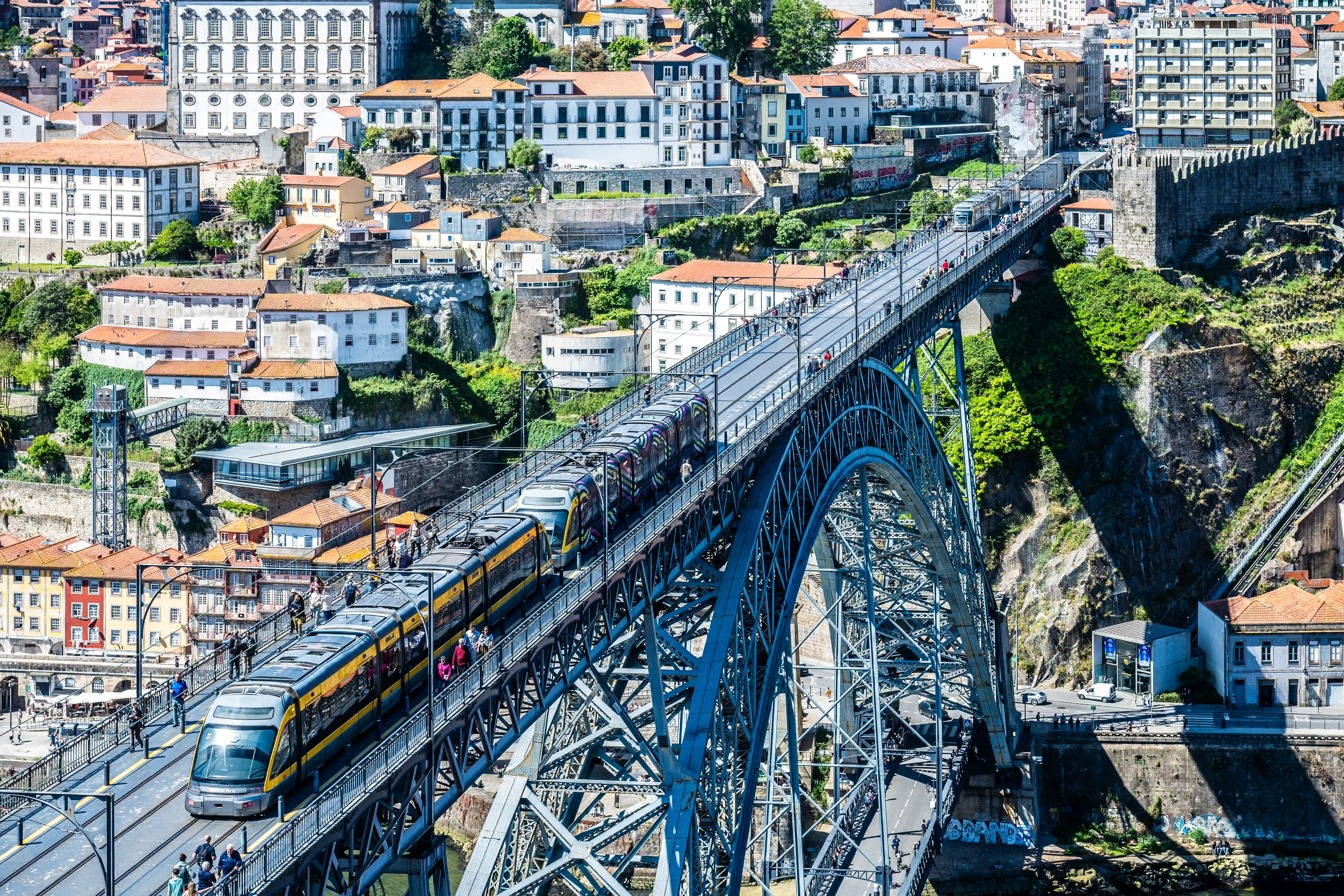  A panoramic view of the Dom Luís I Bridge in Porto, Portugal. A modern tram with yellow and grey stripes, and a graffiti-decorated carriage, moves along the upper level of the metal bridge, alongside numerous pedestrians. The bridge spans a wide river gorge, with a densely built cityscape featuring traditional Portuguese architecture and terracotta rooftops climbing the steep hillsides on both banks. Sunlight creates strong shadows, accentuating the intricate latticework of the bridge and the rugged terrain.