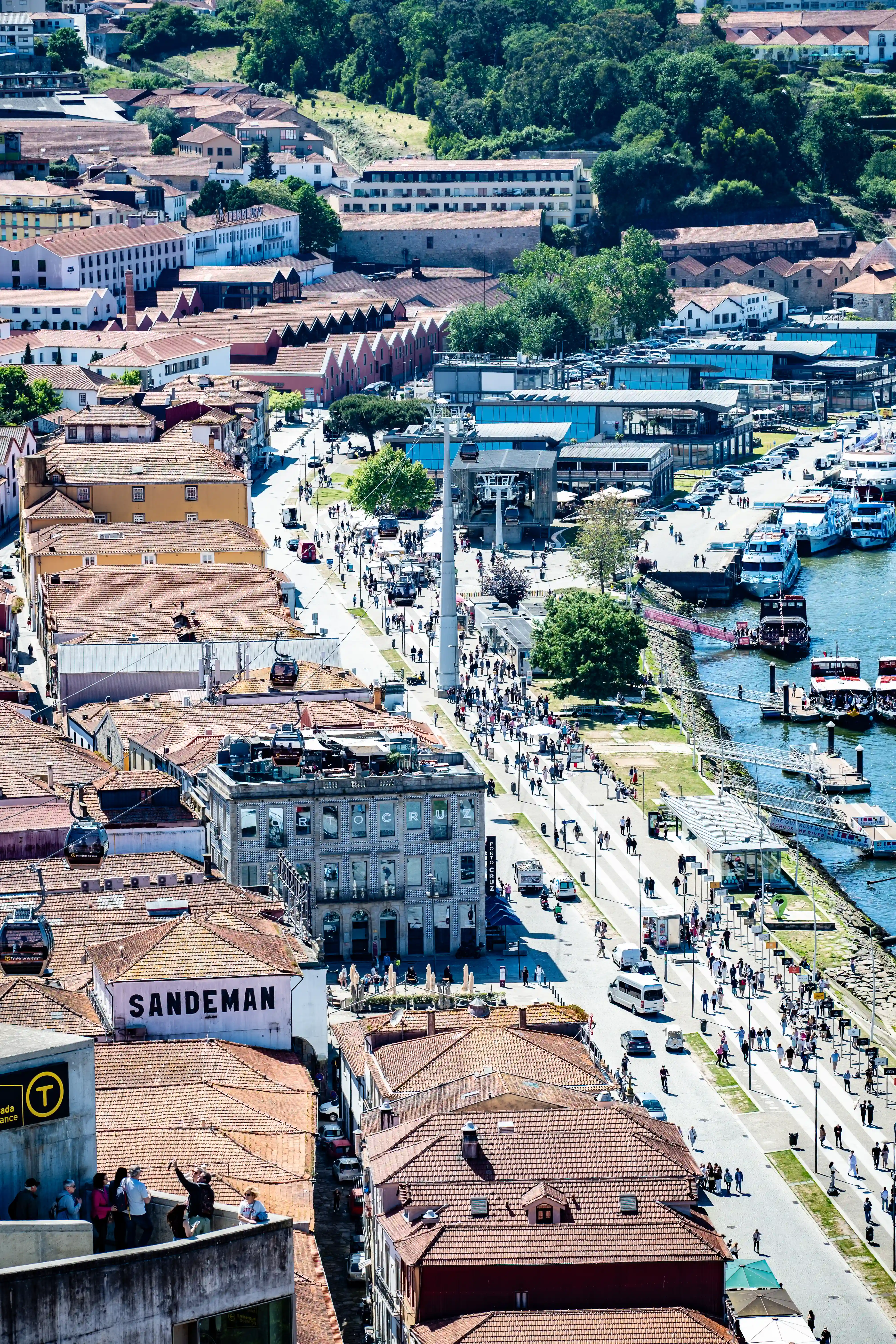 Aerial view of Porto, Portugal, showing a lively riverside promenade with many people walking, a cable car system suspended above, boats docked on the Douro River, and a dense collection of traditional buildings with terracotta roofs.