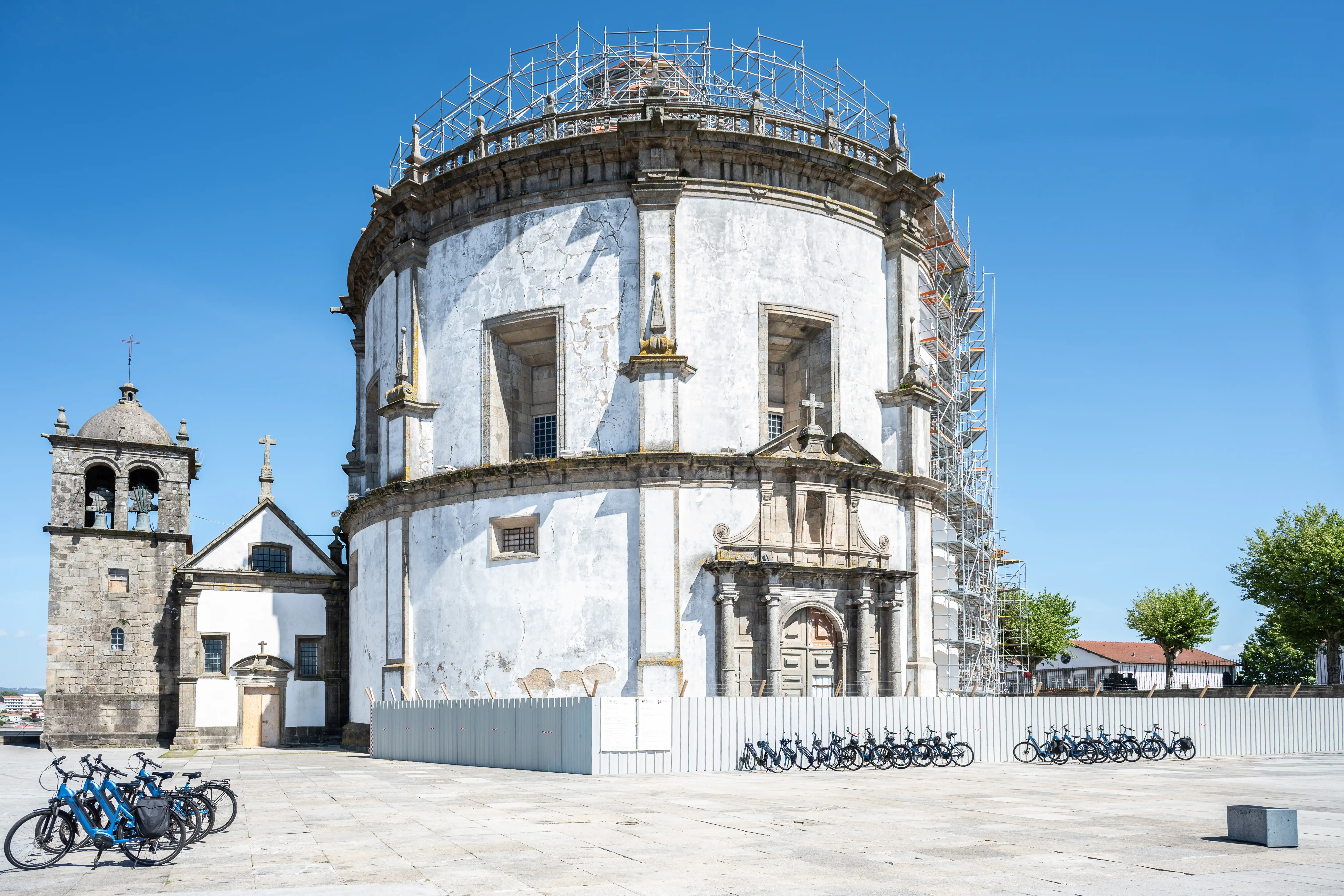 A wide shot of a large, white, circular church under scaffolding, with a stone bell tower to its left, under a bright blue sky. A row of blue rental bicycles is parked in the foreground.
