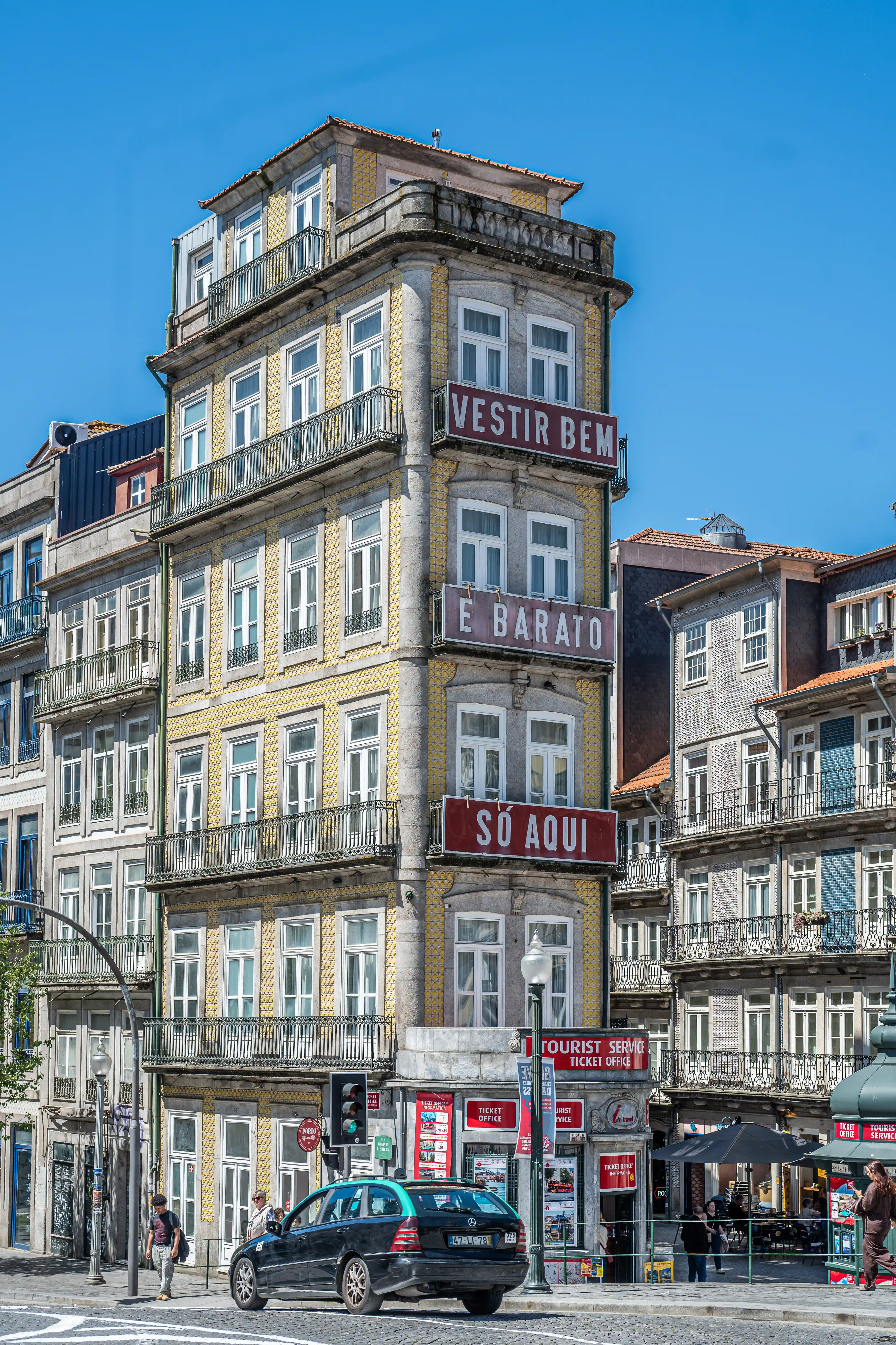 A striking view of a narrow, multi-storey building in Porto, Portugal, adorned with intricate yellow tiles and several balconies. Large red signs with white lettering, VESTIR BEM, E BARATO, and SÓ AQUI, are prominently displayed on the building's facade, indicating a retail establishment. Other buildings with similar traditional Portuguese architectural features, including tiled exteriors and decorative balconies, surround it. A black car is parked on the street below, alongside a building housing a tourist information and ticket office, and people can be seen walking. The image is set against a clear blue sky.