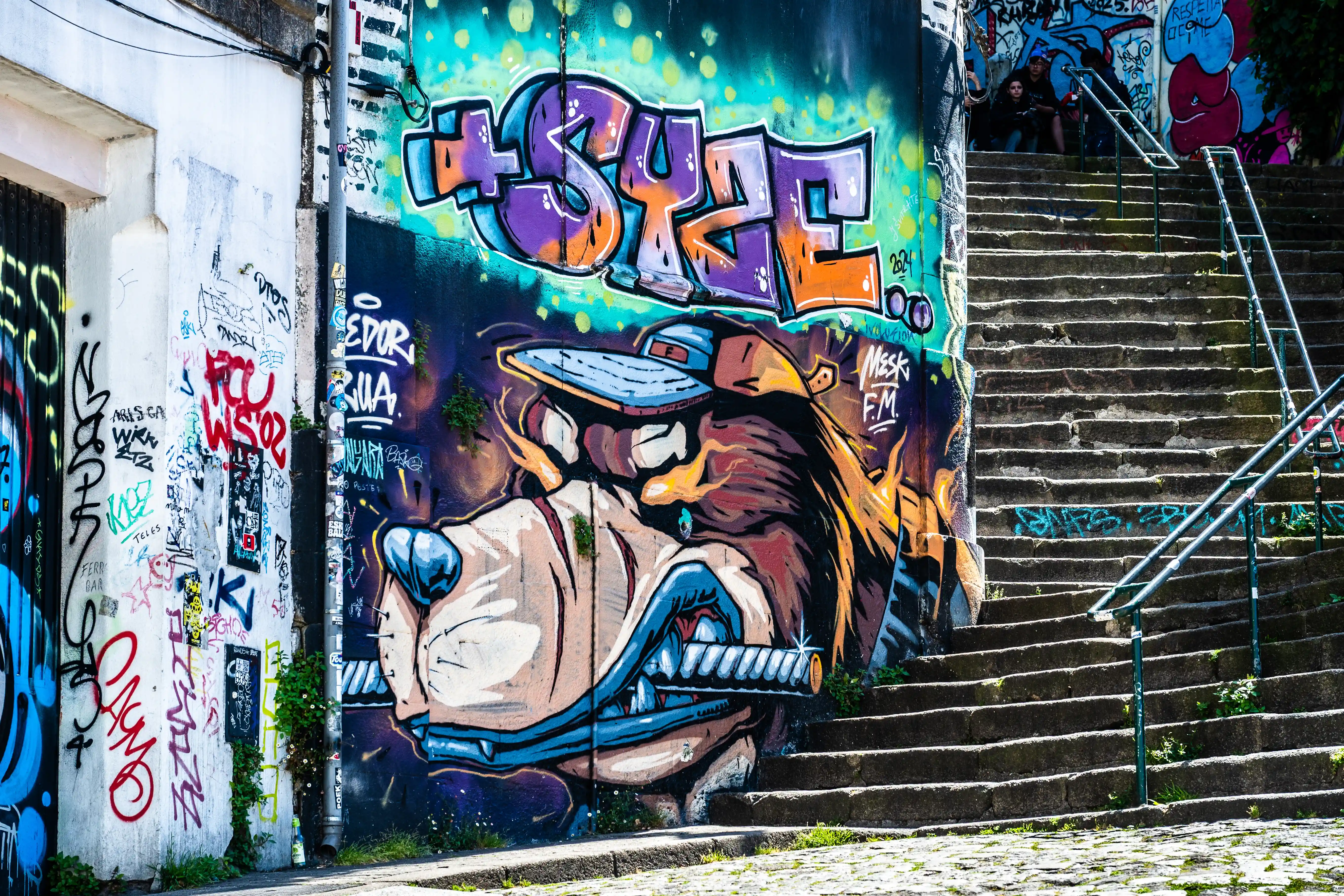 A colourful graffiti mural of a cartoon bear wearing a cap and biting a cylindrical object is painted on a wall next to a stone staircase. The mural features the word 