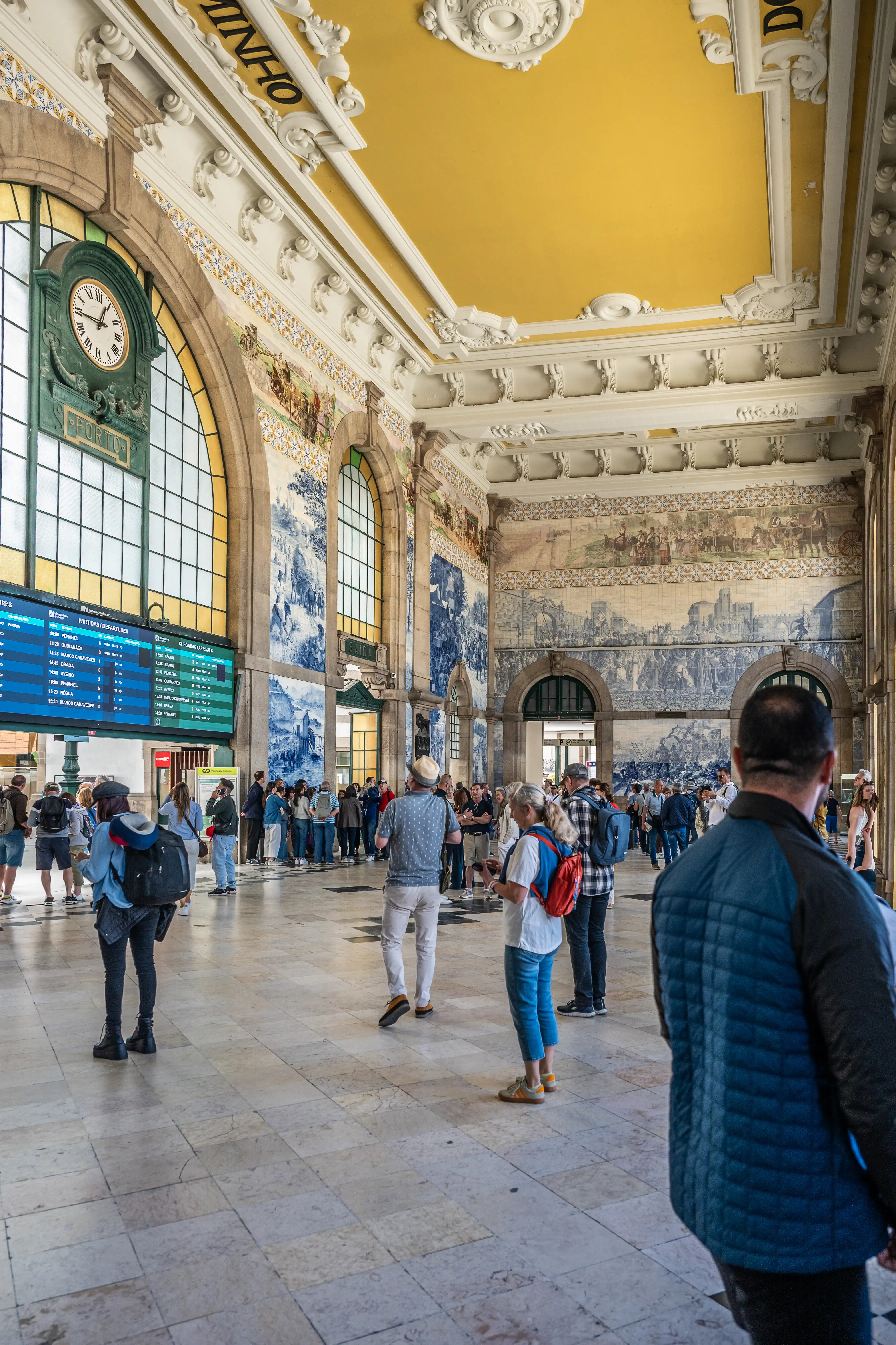 A wide shot of the main hall of São Bento train station in Porto, Portugal, filled with travellers. The walls are covered in intricate blue and white azulejo tiles, depicting various historical scenes. Large arched windows allow natural light to flood the space, highlighting the ornate yellow ceiling and decorative mouldings. A large departure board is visible on the left, and people are seen walking and standing in the hall.