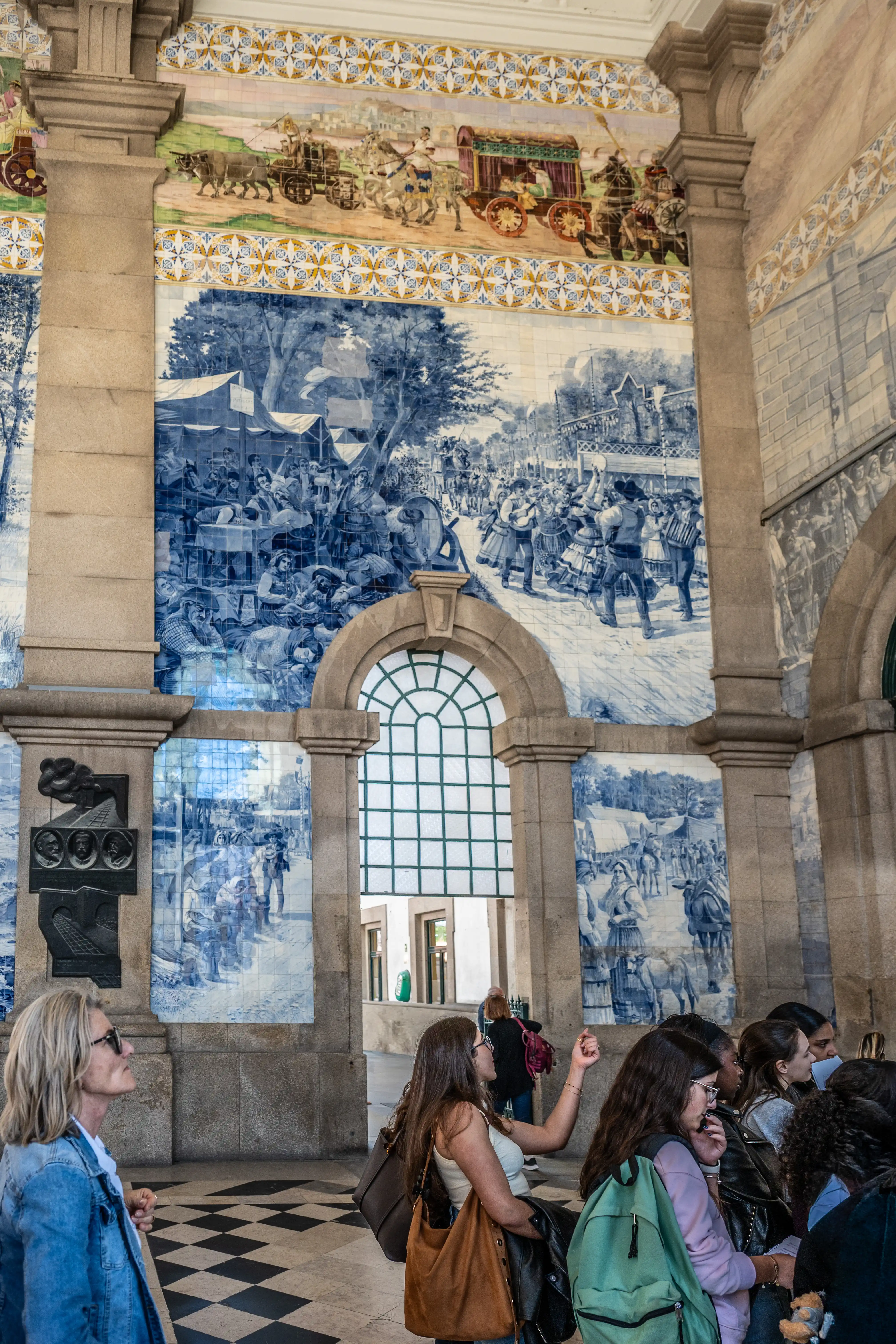 Visitors gaze at large, traditional Portuguese azulejo tiles featuring detailed blue and white historical murals depicting market scenes and transport.