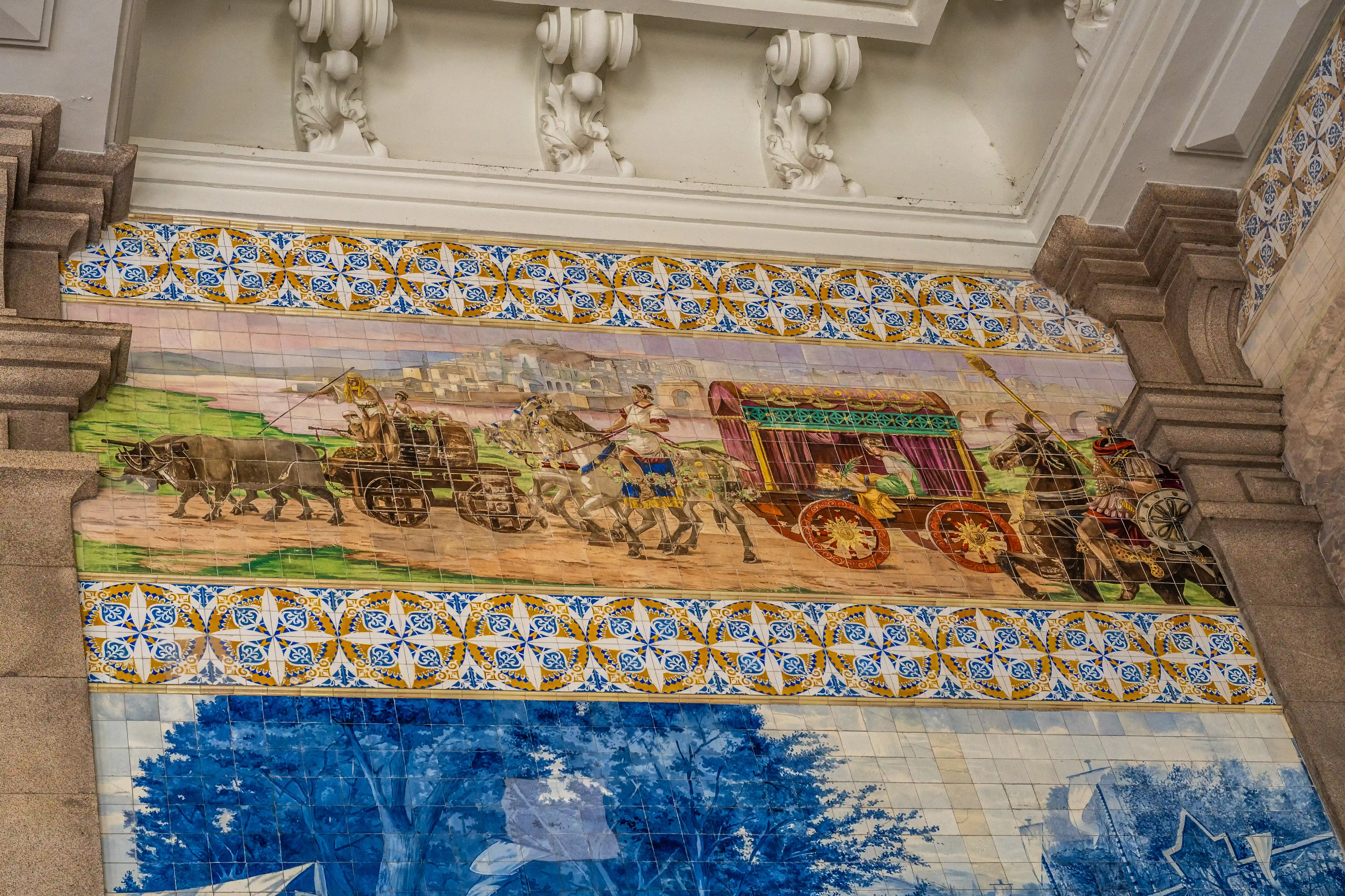  A wide shot of a tiled mural on an exterior wall. The upper section depicts a historical procession with horse-drawn carriages and oxen, bordered by decorative blue and yellow tiles. Below this is a blue and white tiled scene featuring a large tree and buildings.