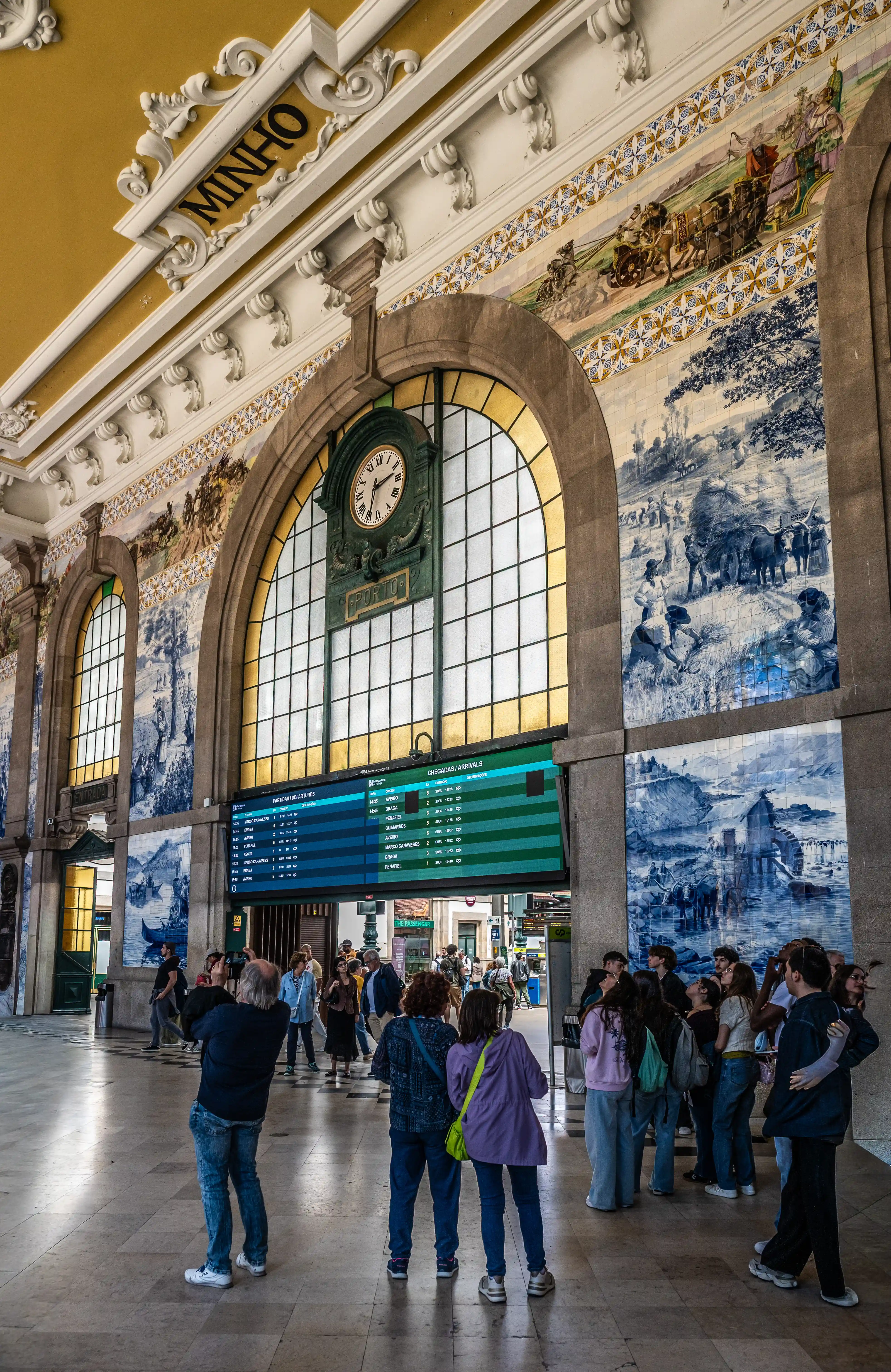 A bustling interior of a grand train station hall adorned with intricate blue and white azulejo tiles, featuring a large digital departures board, a classic clock, and sunlight streaming through arched windows.